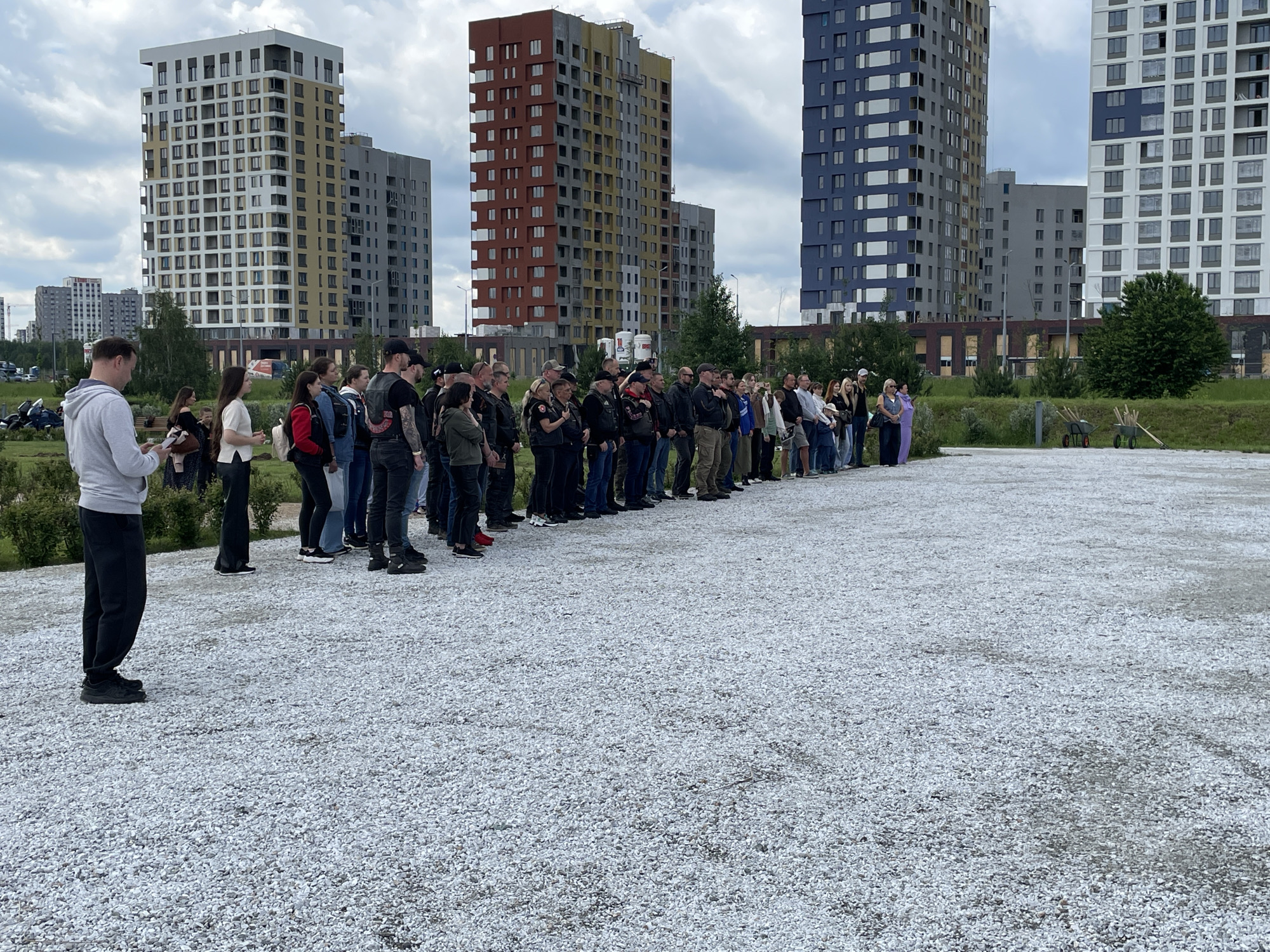 The image depicts an outdoor gathering of approximately 30-40 individuals, primarily adults, standing in a loosely formed line on a light grey gravel surface under an overcast sky. The group is diverse, including men and women of various ages. Several individuals, particularly closer to the left side of the line, are wearing dark vests with patches, indicating affiliation, possibly with a motorcycle club, as several motorcycles are parked in the far left background behind some green shrubs. On the far left, a man in a light grey hoodie and dark pants is visible, looking down at a small electronic device. Towards the far right of the line, two wheelbarrows (one red, one green) and several wooden stakes or tool handles are present on the gravel, suggesting preparation for a planting or landscaping activity.
In the background, a cluster of modern, high-rise residential buildings dominates the skyline. From left to right, the most prominent buildings include:
1. A tall building with white and yellow-beige facade panels.
2. A tall building with reddish-brown, yellow, and grey facade sections.
3. A tall building with dark blue and white facade sections.
Other grey multi-story buildings are visible further in the distance. The general environment is characteristic of a newly developed urban area, with open spaces and contemporary apartment blocks.
The architecture and distinct color schemes of the high-rise buildings in the background identify the location as the **Solnechny (Солнечный) district in Yekaterinburg, Russia**, a known area for modern residential development.
