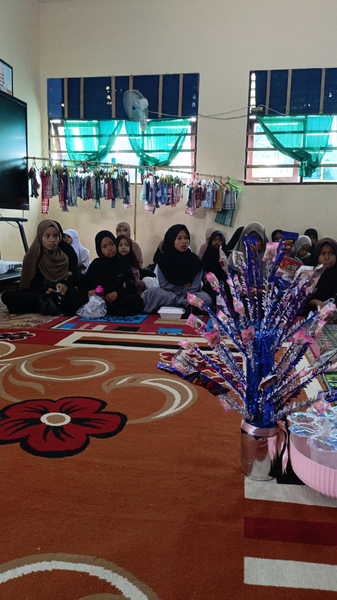 In Saring Sungai Binjai, Indonesia, a group of young girls, dressed in modest Islamic attire, are seated on a patterned orange carpet. The girls are facing forward, suggesting they are engaged in a lesson or presentation. Some of the girls are wearing headscarves in various colors, including brown and black. In the background, two windows with teal curtains are visible, and a fan is mounted on the wall above one of the windows. Clothes are hung on a rack in front of the windows, possibly for a school activity. In the foreground, a decorative tree made of shiny blue and purple material, adorned with small pink and yellow packets, stands in a silver vase. A pink tray holding several clear glasses is placed next to the decorative tree. The room appears to be a classroom or a community hall, with a television on a stand to the left.