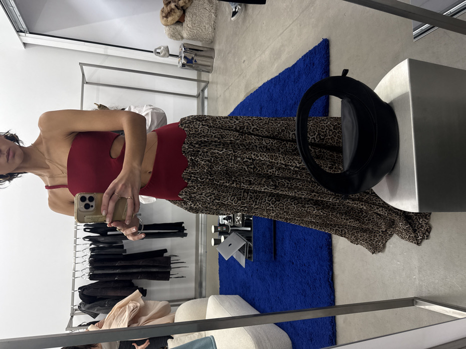 A woman takes a mirror selfie wearing a red, cutout top and a long, leopard print skirt. She's holding her phone, with gold rings on her fingers. The background includes a clothing rack with various garments, a blue rug, and a white sofa. A black handbag rests on a metallic surface. The scene is indoors, likely a store or showroom.