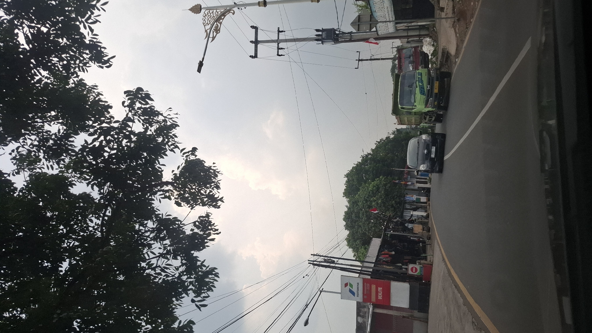 The image depicts a street scene in Sukasari, Indonesia, captured from a vehicle. The perspective is looking forward along a paved road with a yellow line marking the shoulder and a white line dividing lanes. On the left side of the road, there is a red structure with a white sign that reads "PERTAMINA DEXTLITE". Adjacent to this is a collection of stalls or shops with goods displayed outside. Trees line the right side of the road, and further down the road, there are more buildings and utility poles with numerous wires. A black car is driving in the right lane, followed by a green dump truck in the left lane. The sky is overcast with bright clouds. Overhead, dark green foliage from trees partially obscures the sky. In the background, there are glimpses of other vehicles, a transformer box on a utility pole, and decorative elements on a lamppost. The overall environment appears to be a commercial or semi-rural area.