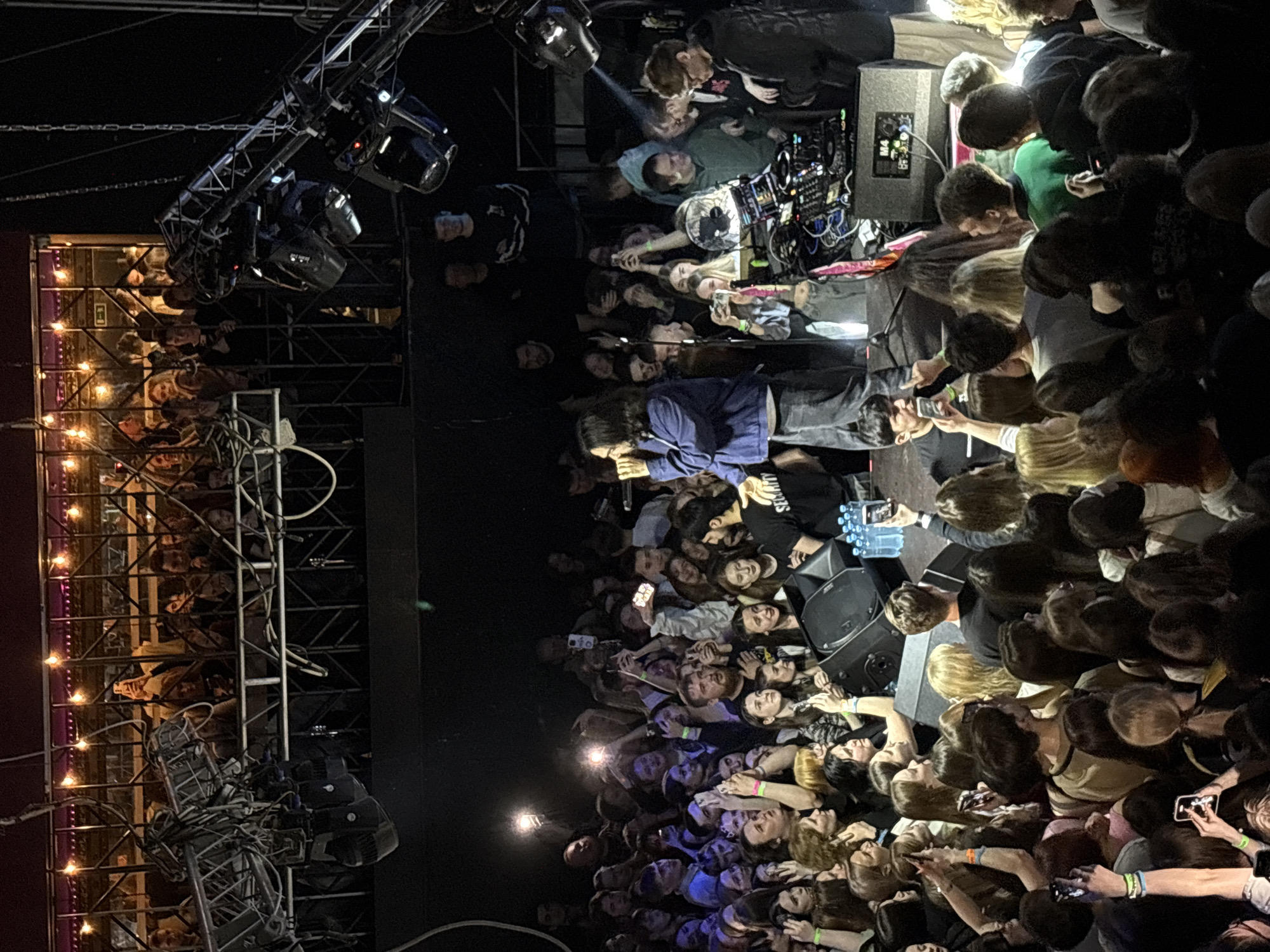 The image depicts a live performance in Izhevsk, Russia. A large crowd of people are densely packed together, facing the stage. Many individuals are holding up their phones, seemingly recording or taking pictures. Onstage, there are speakers, DJ equipment, and lighting rigs overhead. Some people appear to be interacting with the performers, reaching towards them. The lighting is dim, with spotlights illuminating the stage area.