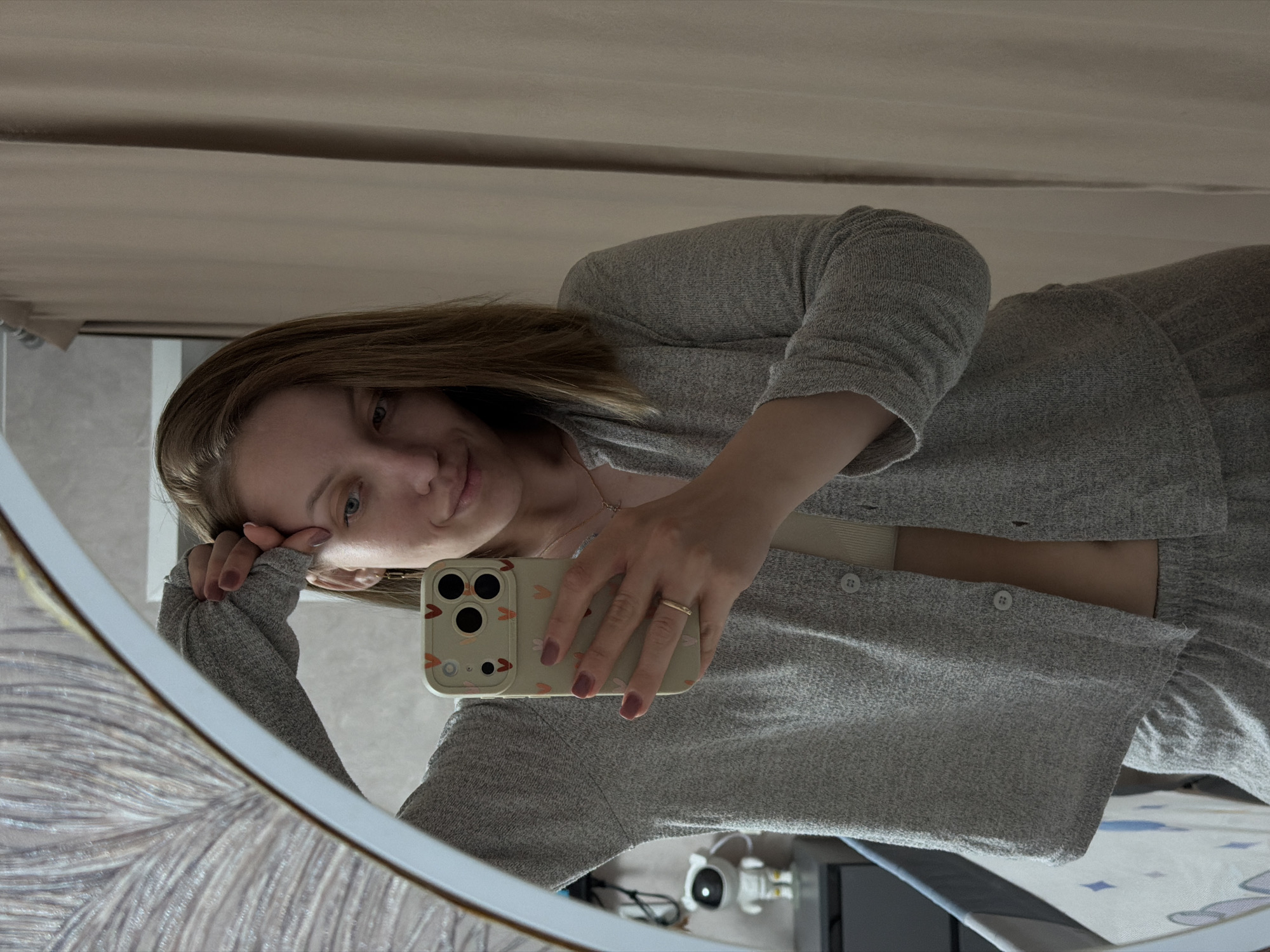 A woman is taking a selfie in a mirror. She is wearing a grey knitted cardigan and matching pants. The background is blurry but appears to be a room with a beige curtain. In the mirror, an ironing board with a patterned cover, eyeglasses, and what appears to be a humidifier are visible. There is no indication of the location being Reshetnikovo, Russia.