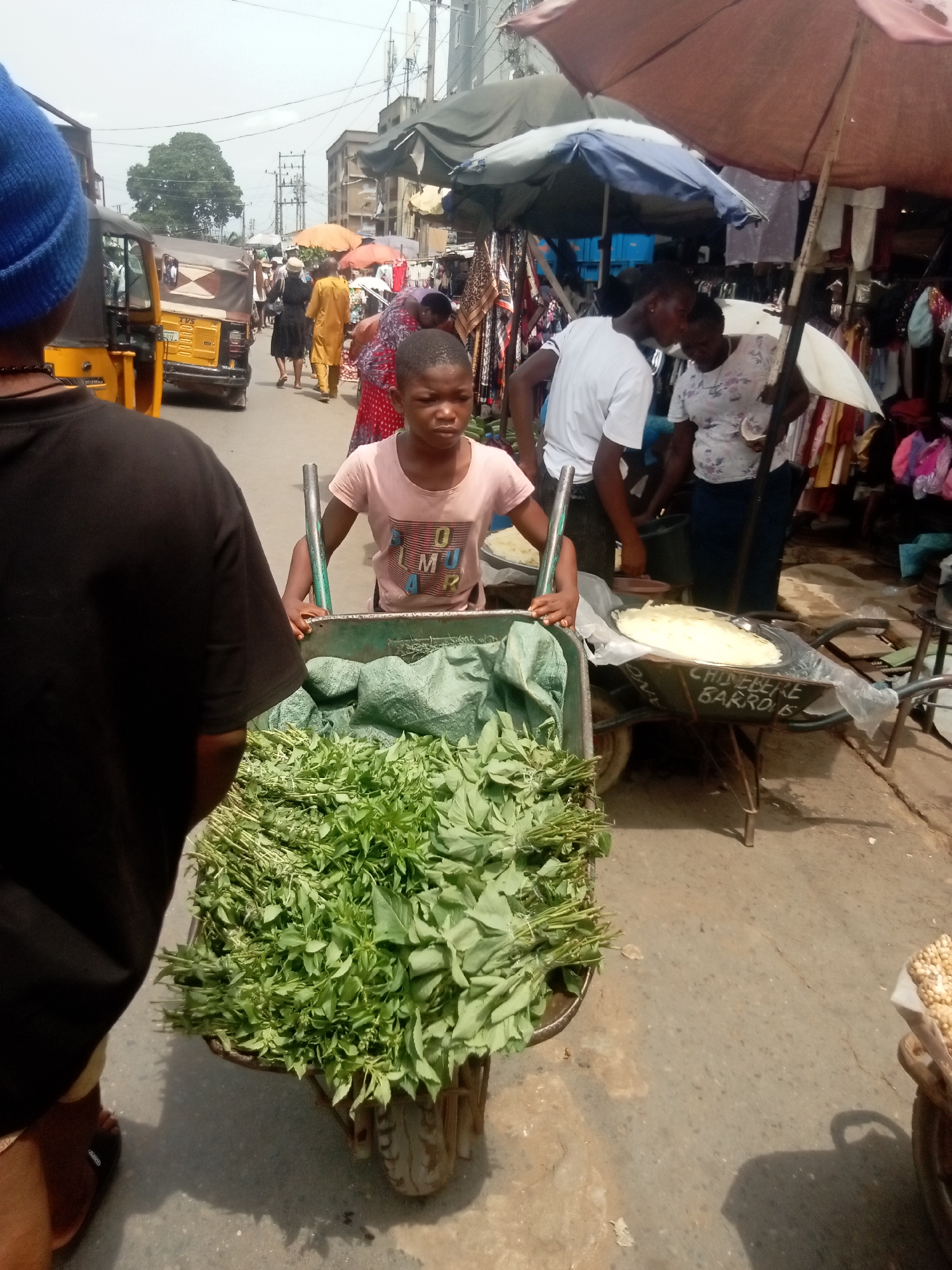 A young person is pushing a wheelbarrow full of leafy green vegetables through a bustling street market in Onitsha, Nigeria. Another wheelbarrow nearby holds a large, flat, white item possibly food. People are shopping and selling under umbrellas and awnings that provide shade from the sun. A yellow auto-rickshaw is visible in the background.