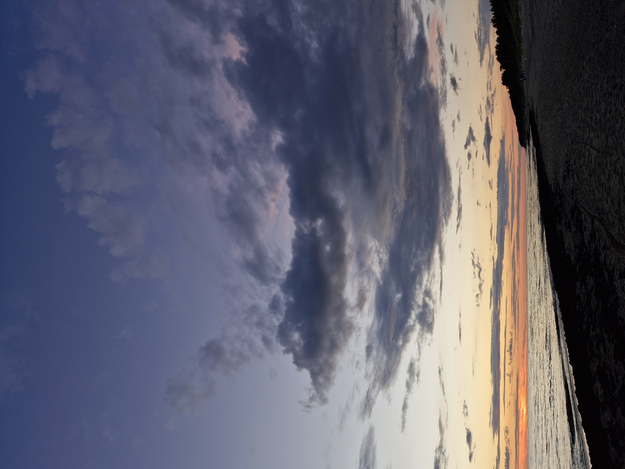 The image depicts a dusk scene at the Curonian Spit, a UNESCO World Heritage site located in Klaipėda, Lithuania. The foreground is a dark, sandy beach with some seaweed or debris scattered near the waterline. The sea extends to the horizon, reflecting the warm hues of the setting sun. The sky above transitions from a soft orange glow near the horizon to a deep blue at the top, with scattered clouds of various shades of gray and purple. A line of dark green trees marks the edge of the coastline in the distance, and a few indistinct figures can be seen walking along the beach.