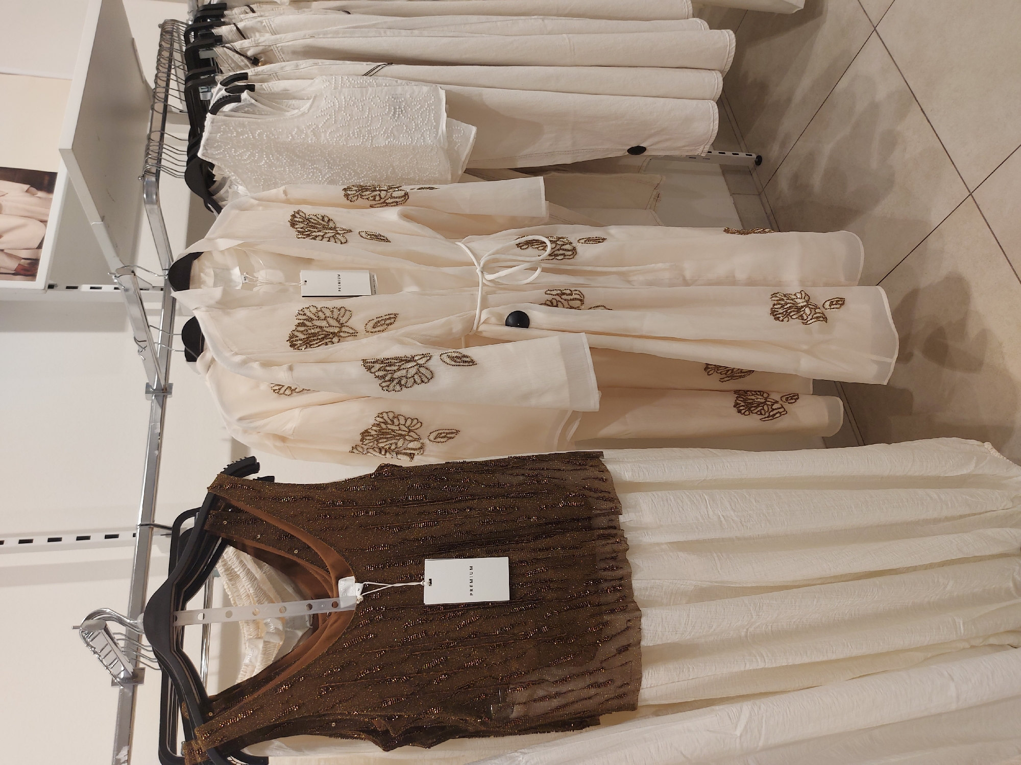 The image shows clothing hanging on a rack in a retail setting. The clothing primarily consists of white and cream-colored garments, some with decorative details. One garment is brown on the top with sequins. All garments are hanging on black hangers. The rack is silver and is placed next to a white wall. The flooring is tiled in light neutral tones. A "Premium" tag is visible on some garments. This scene would have taken place in a clothing store. There is no indication in the image that the location is in Saint Petersburg, Russia.