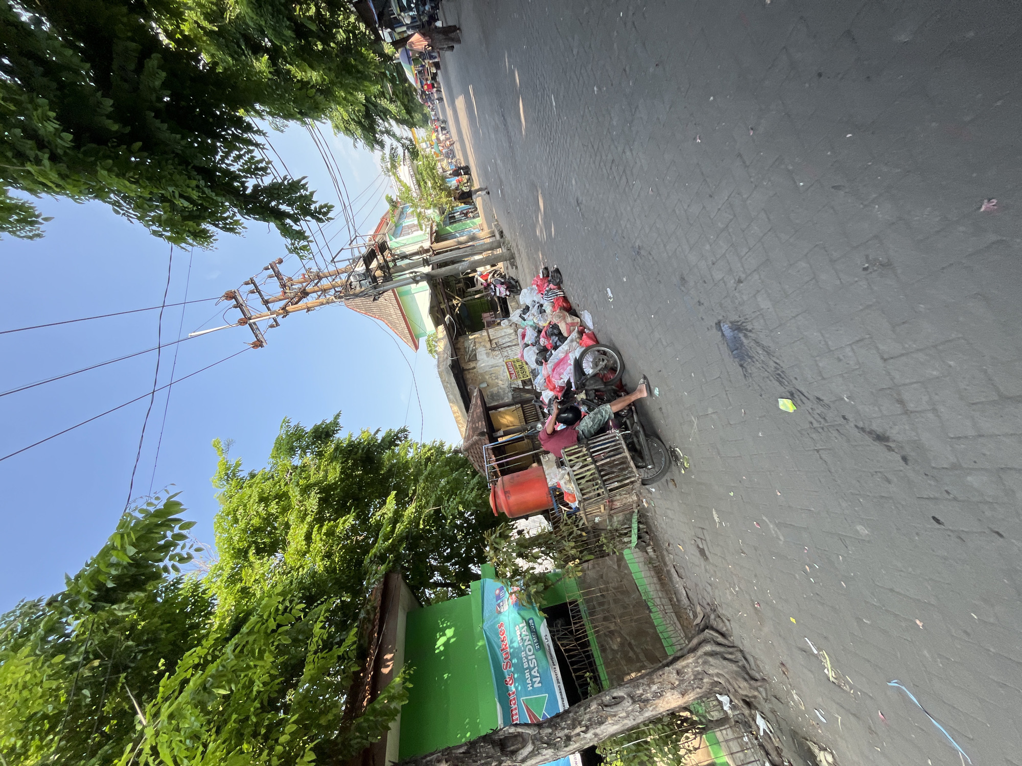 The image depicts a street scene in Juwet Kenongo, Indonesia, with a clear blue sky and lush green trees lining the road. In the foreground, a man is operating a two-wheeled cart loaded with refuse, which is parked next to a collection of garbage bags and assorted items. The cart is equipped with a large red water tank. To the left of the cart, a green building with a banner displaying text in Indonesian can be seen. The street itself is paved with cobblestones, and several motorcycles and a few other people are visible further down the road, suggesting a bustling urban environment. Power lines and a utility pole are prominent in the upper portion of the image. The scene appears to be captured during daylight hours, possibly in the morning or late afternoon, given the quality of light and the shadows.