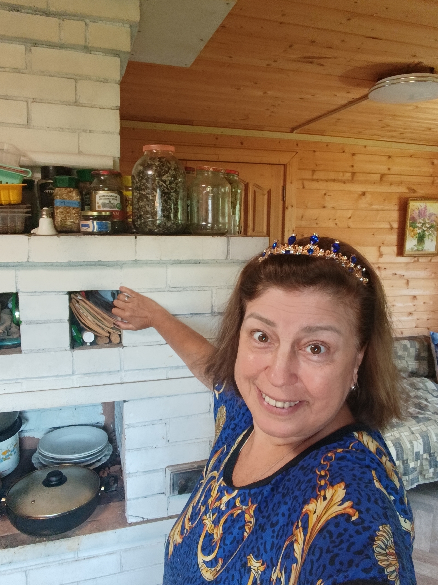 A woman with brown hair and a tiara is standing in front of a white brick structure, possibly a stove or fireplace. She is smiling and pointing towards a small compartment in the brickwork. She is wearing a blue shirt with a gold and black pattern. Various jars and containers are visible on a shelf above the brick structure. The environment appears to be indoors, with wooden walls and ceiling. There is a painting on the wall and a bed in the background.