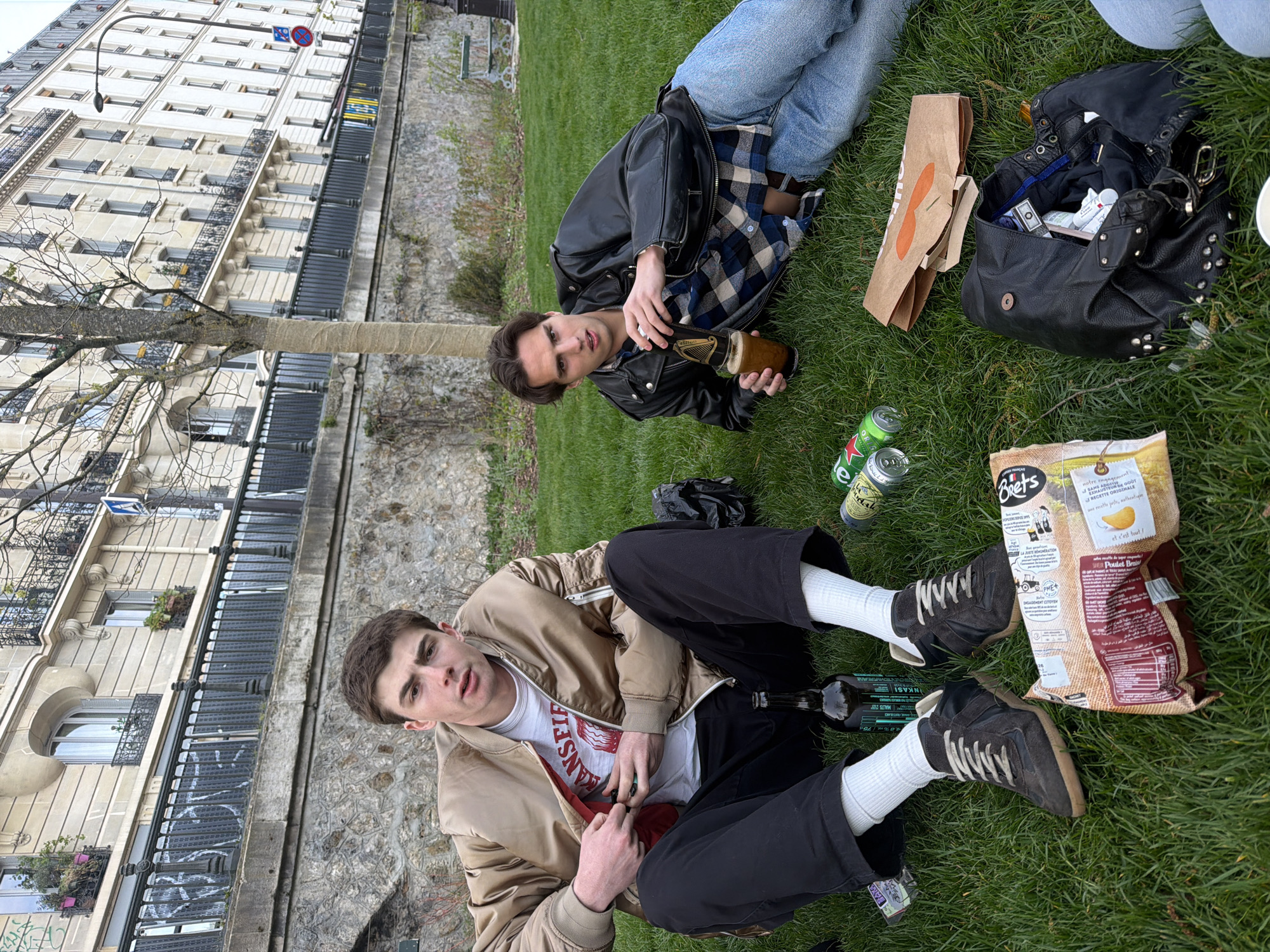 Two men are lying on the grass in a park with a bag of chips, empty beer cans, a black purse, and a takeout food bag scattered around them. The man on the left is wearing a tan jacket over a white t-shirt and black pants with white socks and brown sneakers. He is holding a dark object in his hands, possibly a lighter. The man on the right is wearing a black leather jacket over a blue and black plaid shirt, with light-wash blue jeans. He is holding a brown bottle. The background shows a Parisian building. A tree trunk stands between the two men and the building. The image is rotated. The image is likely taken in Paris, France, based on the architectural style of the building.
