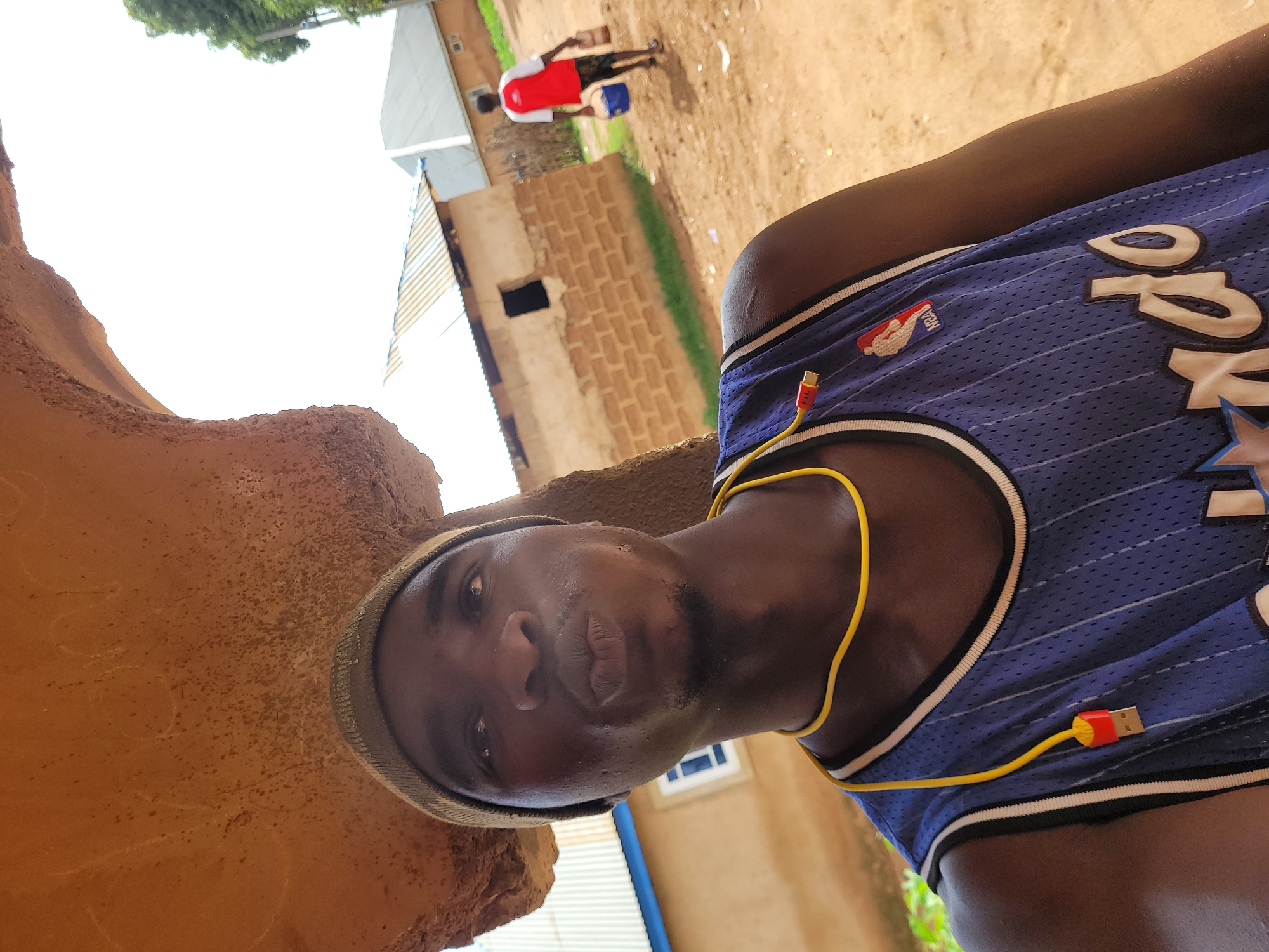 In Bukuru, Nigeria, a young man wearing a green beanie and a blue basketball jersey with "Orlando" written on it poses for a selfie. He has a yellow cable with a red USB connector hanging around his neck, with the connector clipped to his jersey. Behind him, a man in a red and white shirt carries two buckets, walking on a dirt path towards brick buildings with corrugated metal roofs. The scene is outdoors under a bright sky, with some greenery visible on the right side of the frame.