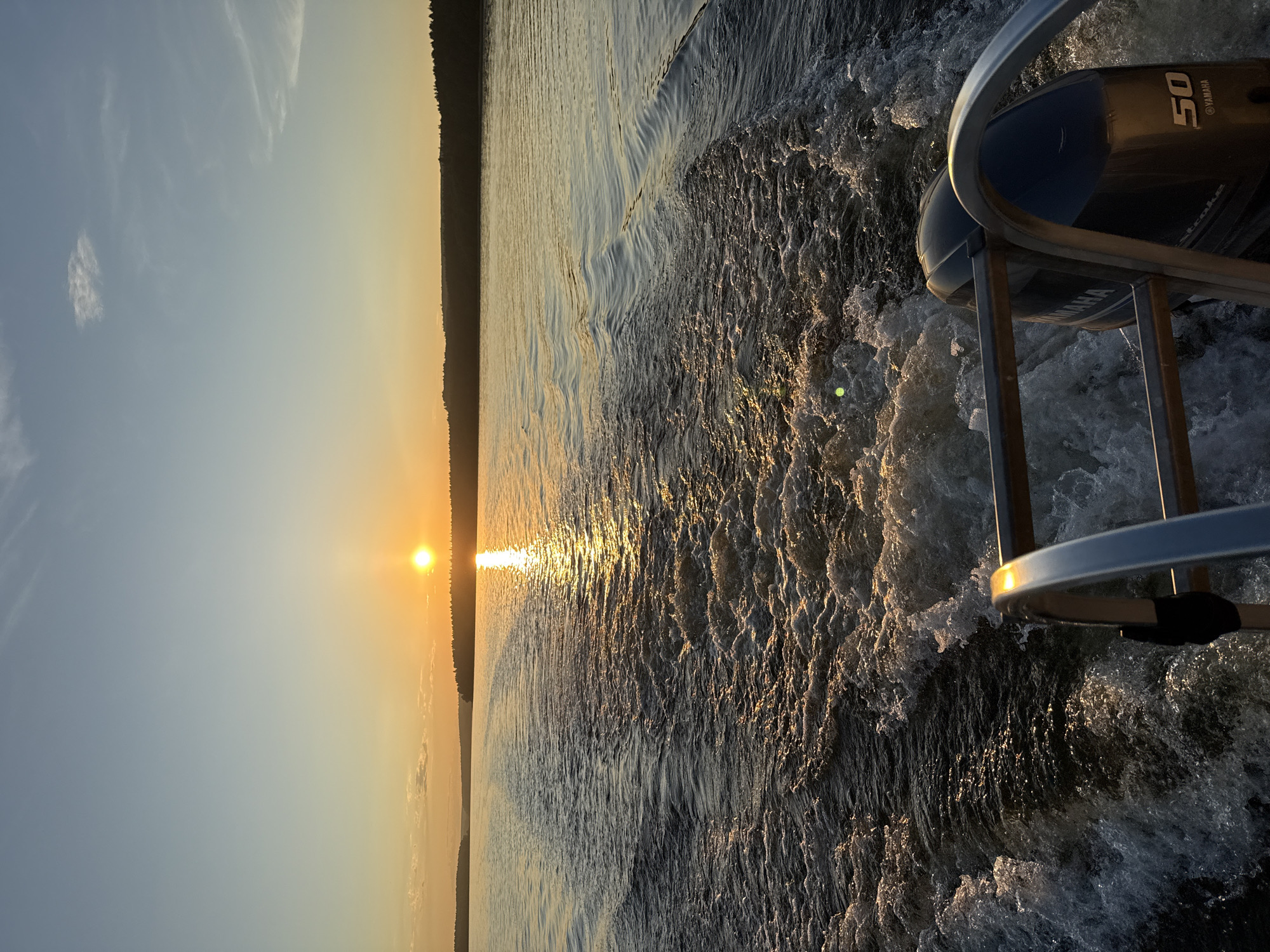 The image captures a sunset from the stern of a moving boat on a body of water near Votkinsk, Russia. A prominent, bright orange sun is positioned low on the horizon, casting a strong, shimmering reflection that extends across the water towards the viewer. The sky exhibits a gradient, transitioning from a light blue at the top to warm orange and yellow hues near the horizon, with a few faint, wispy clouds visible.
The midground features relatively calm water with subtle ripples, while the immediate foreground is dominated by a turbulent, churning white wake, indicative of the boat's forward motion. A dark, forested landmass forms the distant horizon line on both sides of the water. In the lower right of the frame, the upper casings of two dark outboard motors are visible, with the one on the right clearly displaying "YAMAHA" in white lettering, followed by "4 Stroke" and the number "50," likely denoting horsepower. Silver-colored metal railings, part of the boat's structure, are prominently positioned in the foreground, framing the view of the wake and motors. No people are visible in the scene.