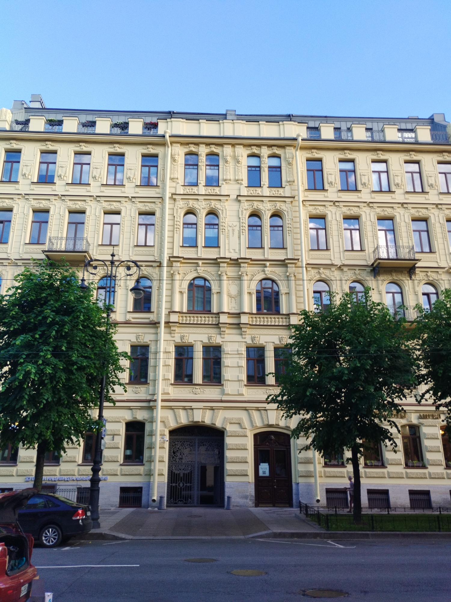 This image depicts the facade of a multi-story building in Saint Petersburg, Russia. The building is painted a pale yellow and features ornate architectural details, including decorative moldings, pilasters, and arched windows on the upper floors. The ground floor has large, dark wooden double doors with intricate metal gates in front of the entryway. Several trees with lush green foliage are situated in front of the building, and a street lamp with a decorative design stands to the left. A dark blue sedan is parked on the street, and a red car is partially visible in the foreground. The sky is clear and blue. The scene appears to be a daytime shot on a street in a European city.