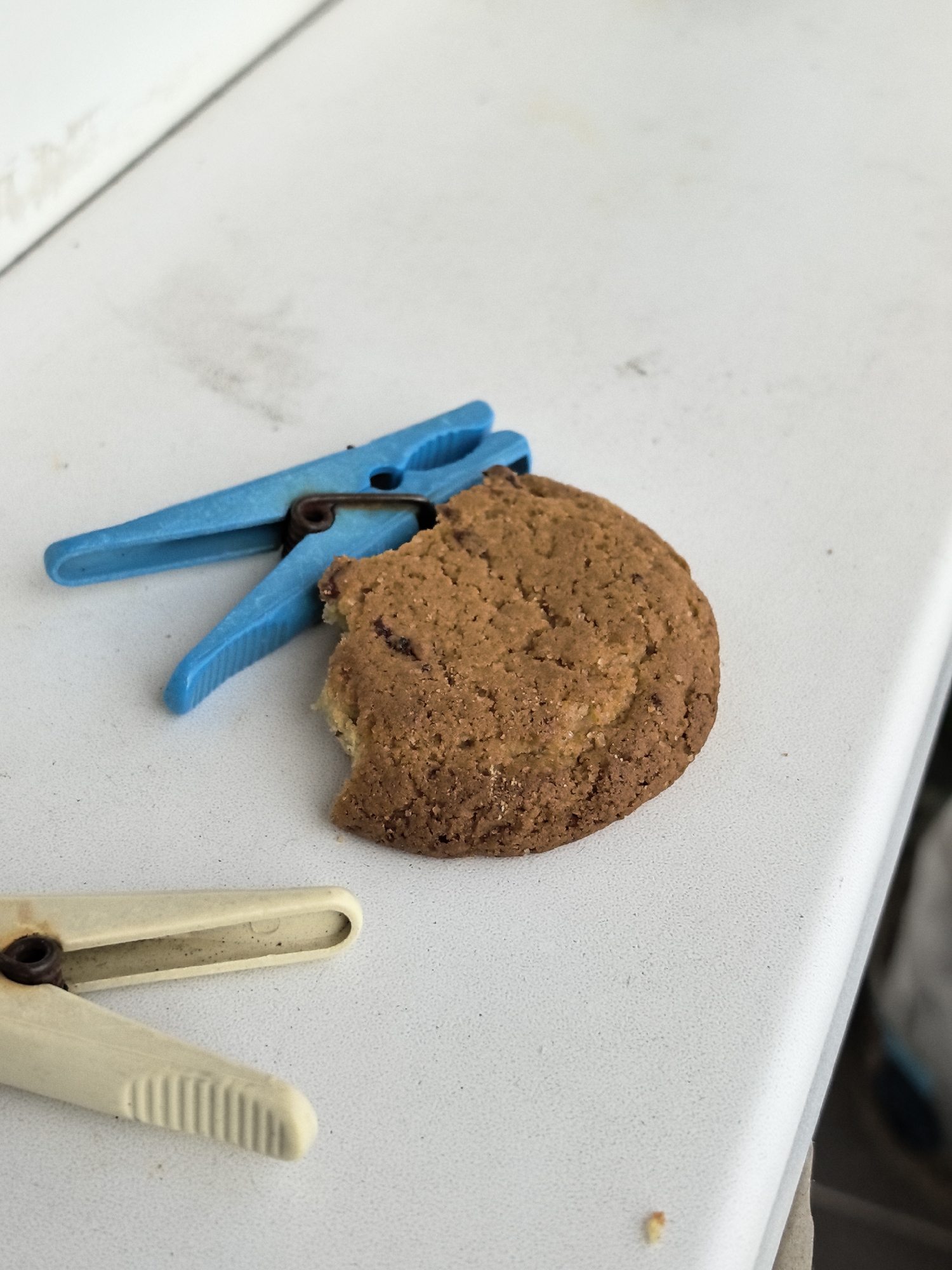 This image shows a close-up of a cookie with a bite taken out of it, placed on a white surface. Two clothespins are also visible in the frame: one is blue and positioned to the left of the cookie, and the other is beige and located in the bottom left corner. The surface appears to be a countertop or a similar flat surface, with some light smudges and dust. The lighting suggests it might be daytime, with the light source coming from the top. The overall scene is simple and domestic, with no people or explicit text visible. The focus is on the cookie and the surrounding objects.