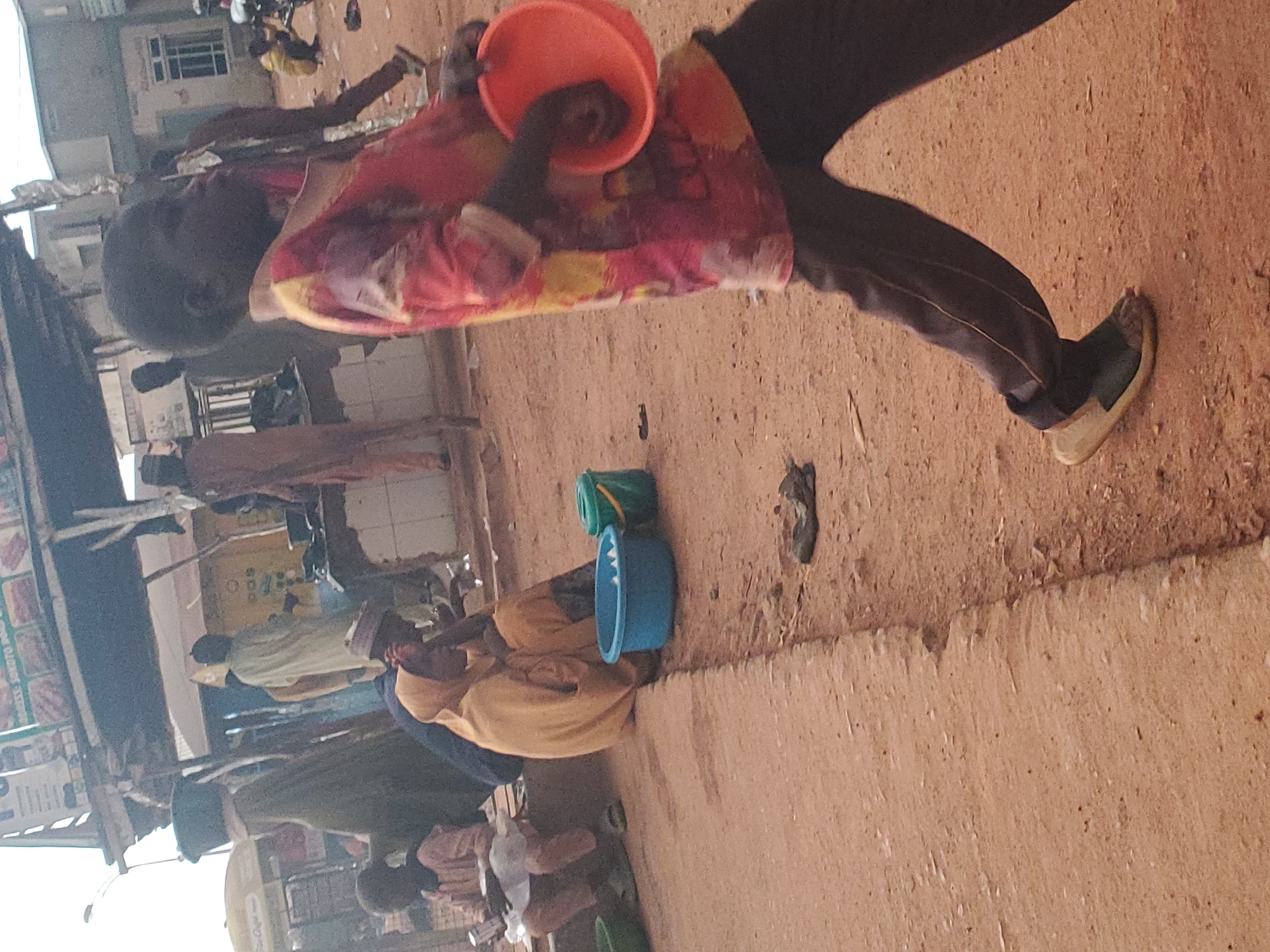 This is a daytime, full-shot image depicting a street scene in Funtua, Nigeria. The foreground is dominated by a young Black boy walking from right to left, wearing a vibrant, patterned long-sleeved shirt and dark trousers. He holds an orange bowl in his right hand. To the left of the boy, a woman wearing a yellow hijab and gown sits on a curb, with her back to the camera. Next to her are two buckets, one blue and one green, on the ground. Further back, other individuals are visible, some standing and some sitting, near market stalls. The environment appears to be a dusty, unpaved street with a building structure providing shade behind the market stalls. The overall impression is one of a busy, everyday urban environment.