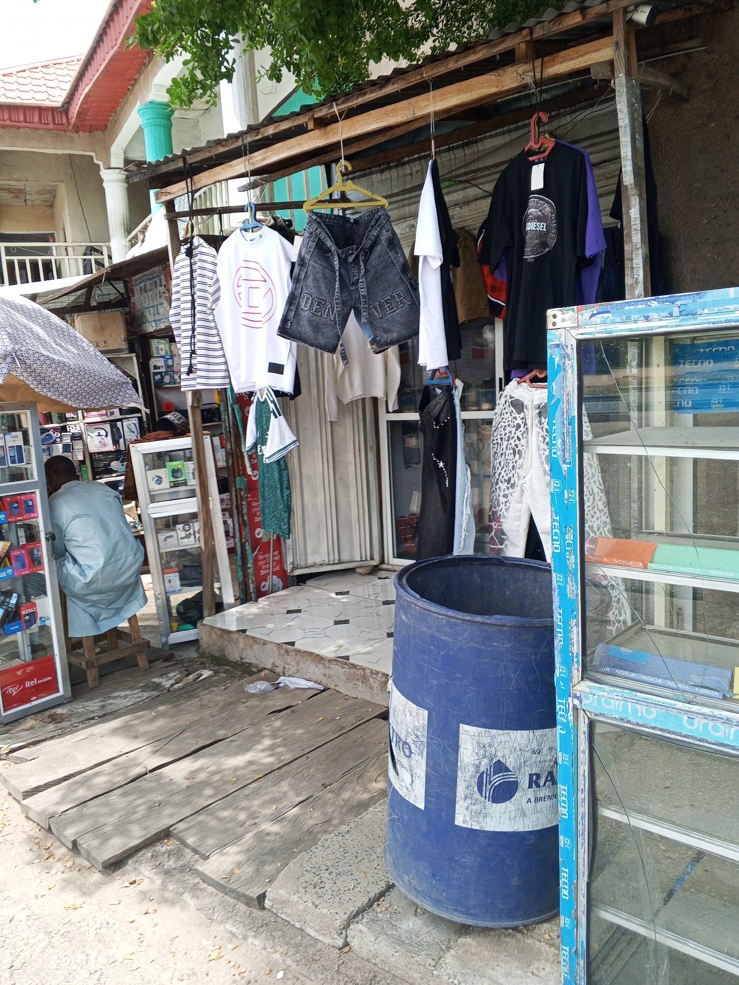 The image depicts an outdoor market stall in Jalingo, Nigeria. A person is seated on a wooden stool inside a stall filled with electronic devices, likely phones and accessories, displayed in glass cabinets. Outside, various clothing items, including t-shirts and shorts, are hanging on display racks. A large blue plastic barrel stands in front of the stall, and a glass display cabinet is positioned to the right. Wooden planks form a makeshift pathway in front of the stall. The surrounding environment includes building structures and trees, indicating a typical urban setting in Jalingo.