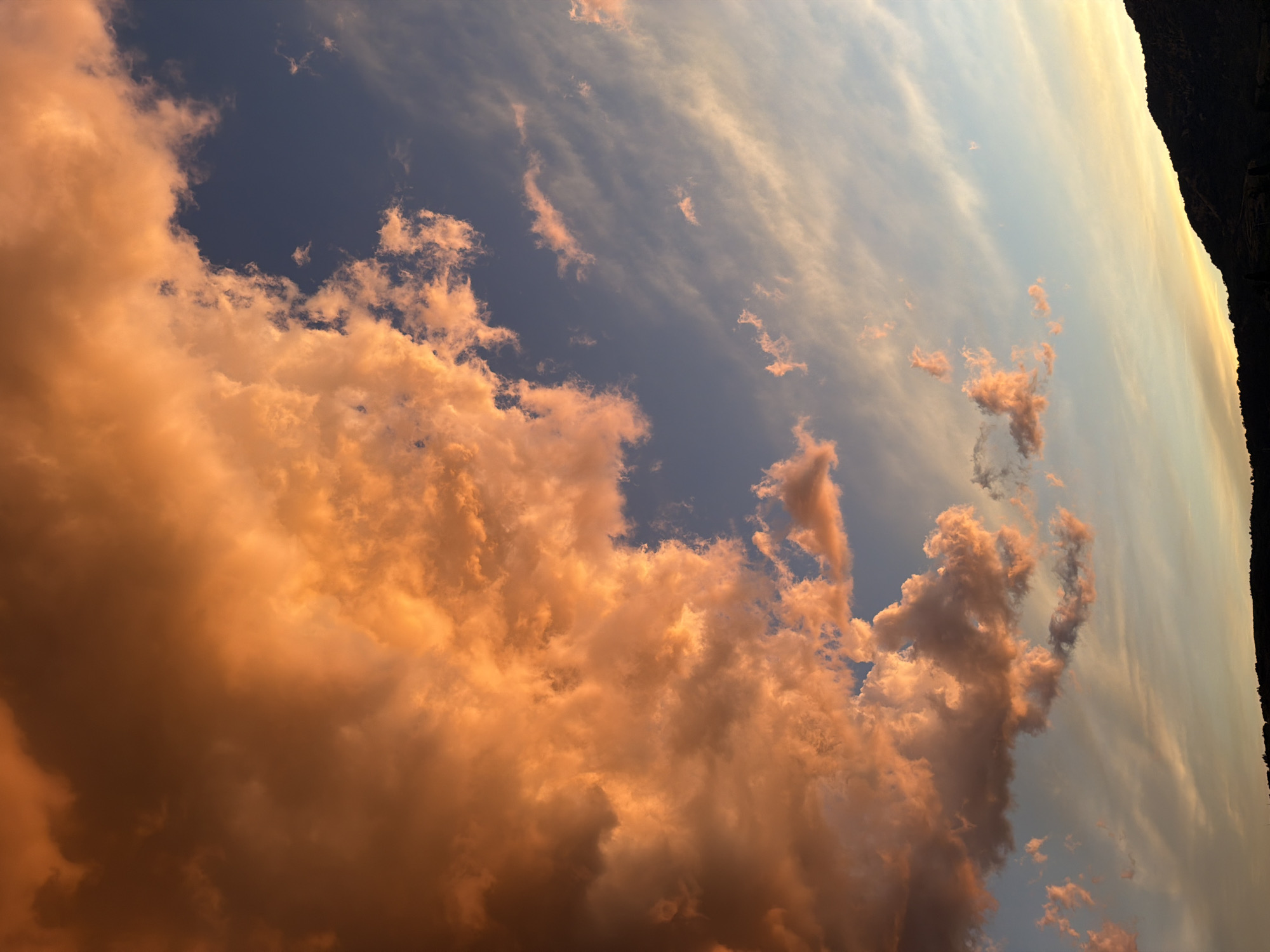 A dramatic sunset sky over Guilherand-Granges, France, is filled with large, fiery orange clouds that dominate the left side of the frame. These clouds are illuminated by the setting sun, creating a vibrant and textured appearance. To the right, the sky transitions to a softer blue, with wispy, lighter clouds streaking across it, reflecting the fading light. The lower portion of the image shows a dark silhouette of rolling hills or mountains, with the very top edge of a distant town or settlement barely visible against the horizon. The scene captures the intense colors and dramatic formations of clouds during the golden hour.
