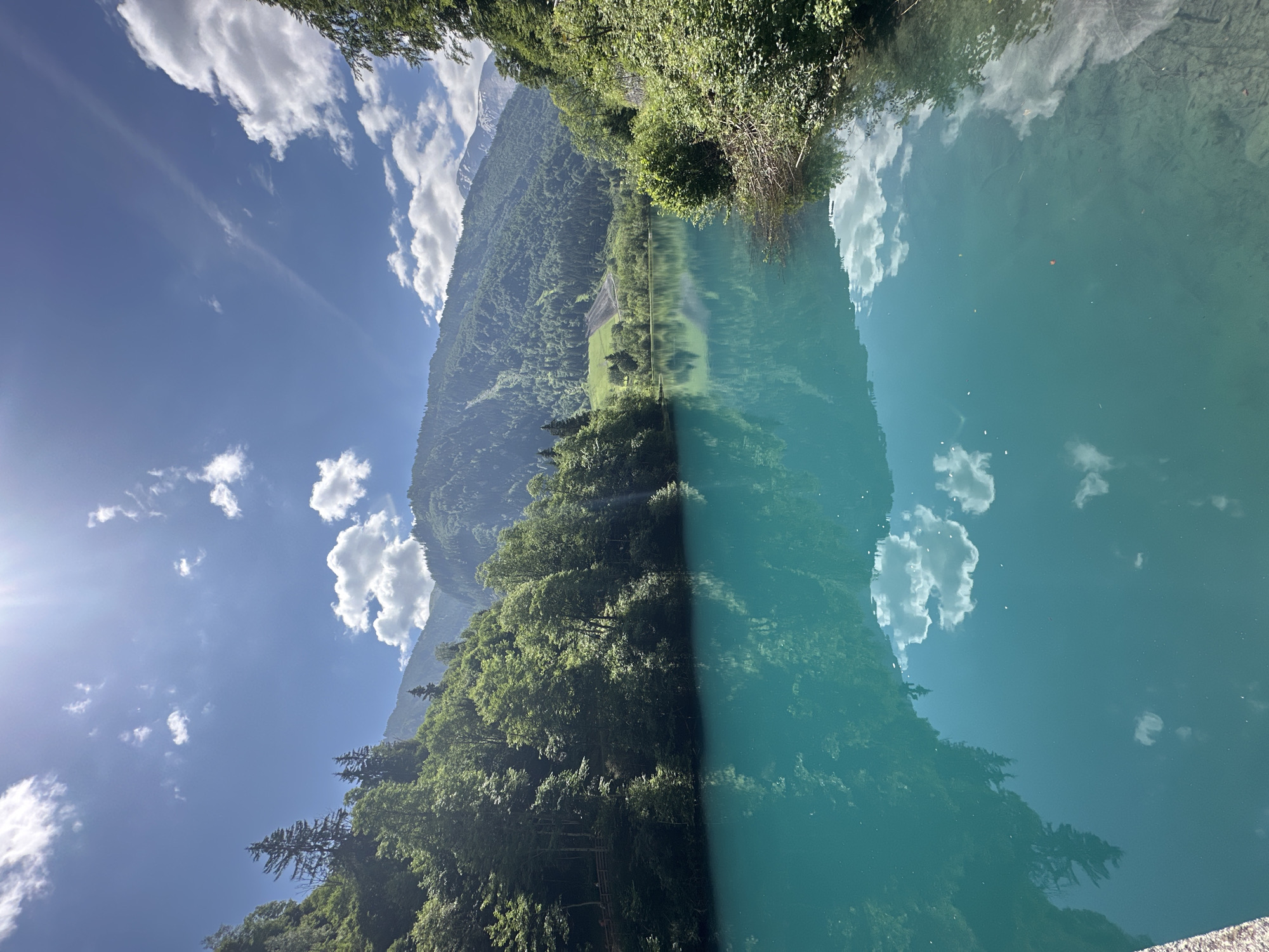 This media file depicts a serene, vertical landscape scene of a body of water reflecting the surrounding environment. The image appears to be taken in Kaprun, Austria.
The primary subjects are the crystal-clear, turquoise water occupying the right half of the frame, and the lush, green mountainside and forest that dominate the left half. The mountains are covered in dense vegetation, with some patches of lighter green suggesting clearings or meadows. The sky above is a vibrant blue, dotted with fluffy white clouds, some of which are also reflected in the water. A bright sunbeam pierces through the clouds on the left side of the image, creating a lens flare effect. The reflection in the water is remarkably clear, showing an upside-down image of the mountains, trees, and sky.
No people or specific objects are clearly discernible, though the reflections hint at the presence of trees and the natural landscape. The overall impression is one of tranquility and natural beauty. The lighting suggests it is daytime, likely mid-morning or afternoon, given the bright sun and clear skies. The weather appears to be fair and sunny.
There is no visible text in the image. The location cues, such as the mountainous terrain and the distinct color of the water, are consistent with alpine regions like Kaprun, Austria. The scene evokes a sense of peace and awe at the power and beauty of nature.