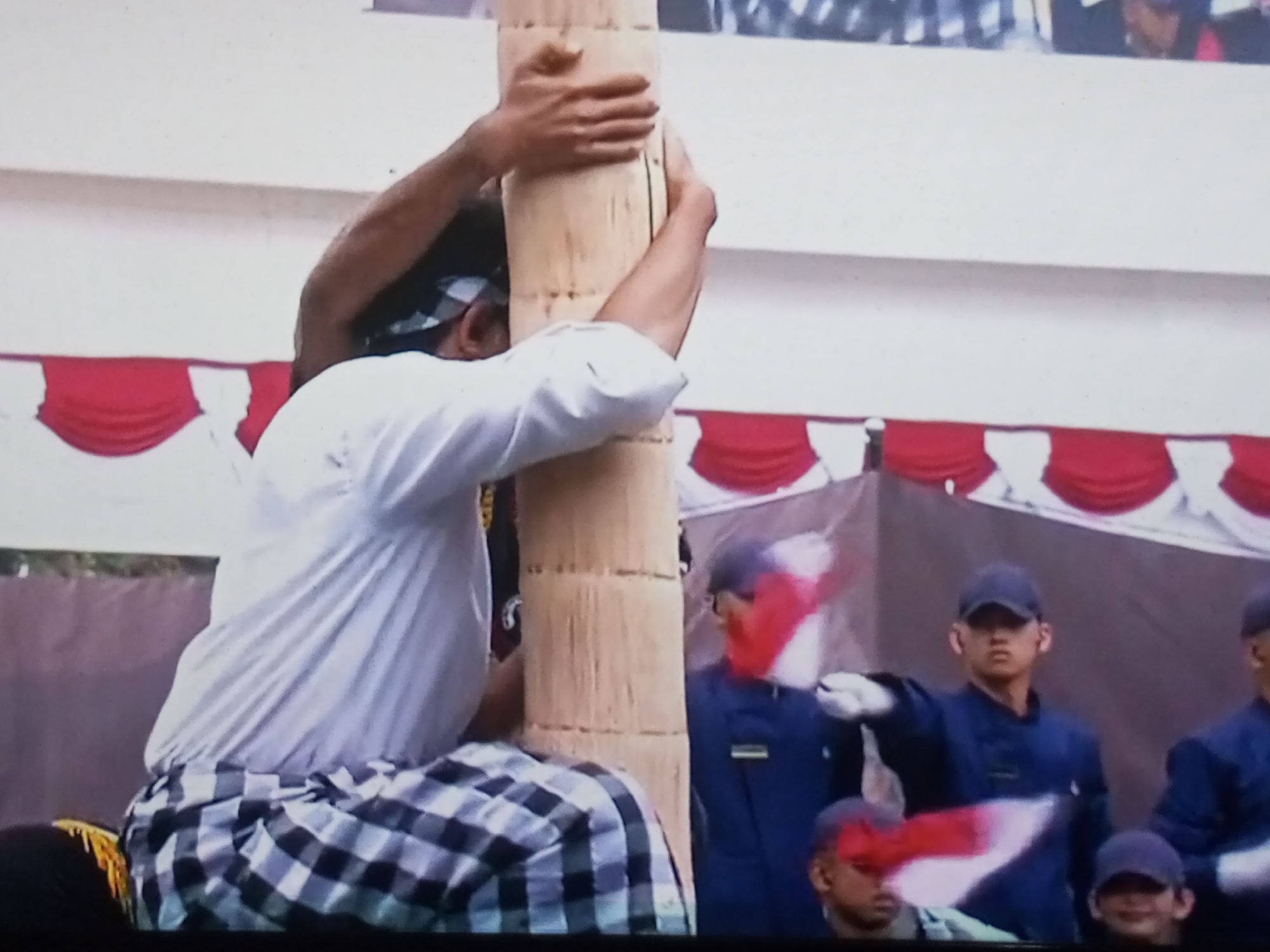 In Banyudono, Indonesia, a man in a white shirt and a checkered sarong is climbing a wooden pole. His arms are wrapped around the pole, and he is looking downwards. Behind him, several people in blue uniforms and caps are visible, some holding Indonesian flags. The background features red and white decorative banners.