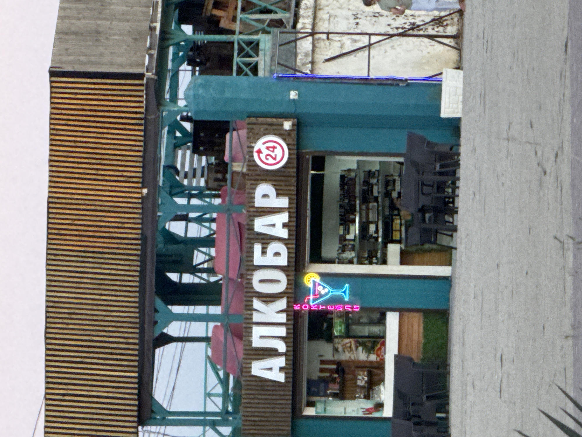 The image shows a facade of a bar or establishment in Novofedorovka, Ukraine. A prominent sign displays the Cyrillic word "АЛКОБАР" in white, backlit letters, indicating an "Alcohol Bar." To the right of the word, there is a red circular sign with "24" inside, suggesting it is open 24 hours. Below the main sign, a neon sign in blue features a martini glass with a lime and an olive, and the word "КОКТЕЙЛИ" (cocktails) in pink vertical lettering. The establishment has large windows, revealing shelves stocked with bottles and other items inside. In front of the bar, several dark-colored tables and chairs are set up on a light-colored paved area. A turquoise-colored pillar is visible to the right of the bar's entrance. In the background, there is a structure with wooden slats and a green railing. A person, partially visible on the far right, is wearing light-colored pants and a green t-shirt. The overall environment appears to be an outdoor seating area of a commercial establishment in a public space.