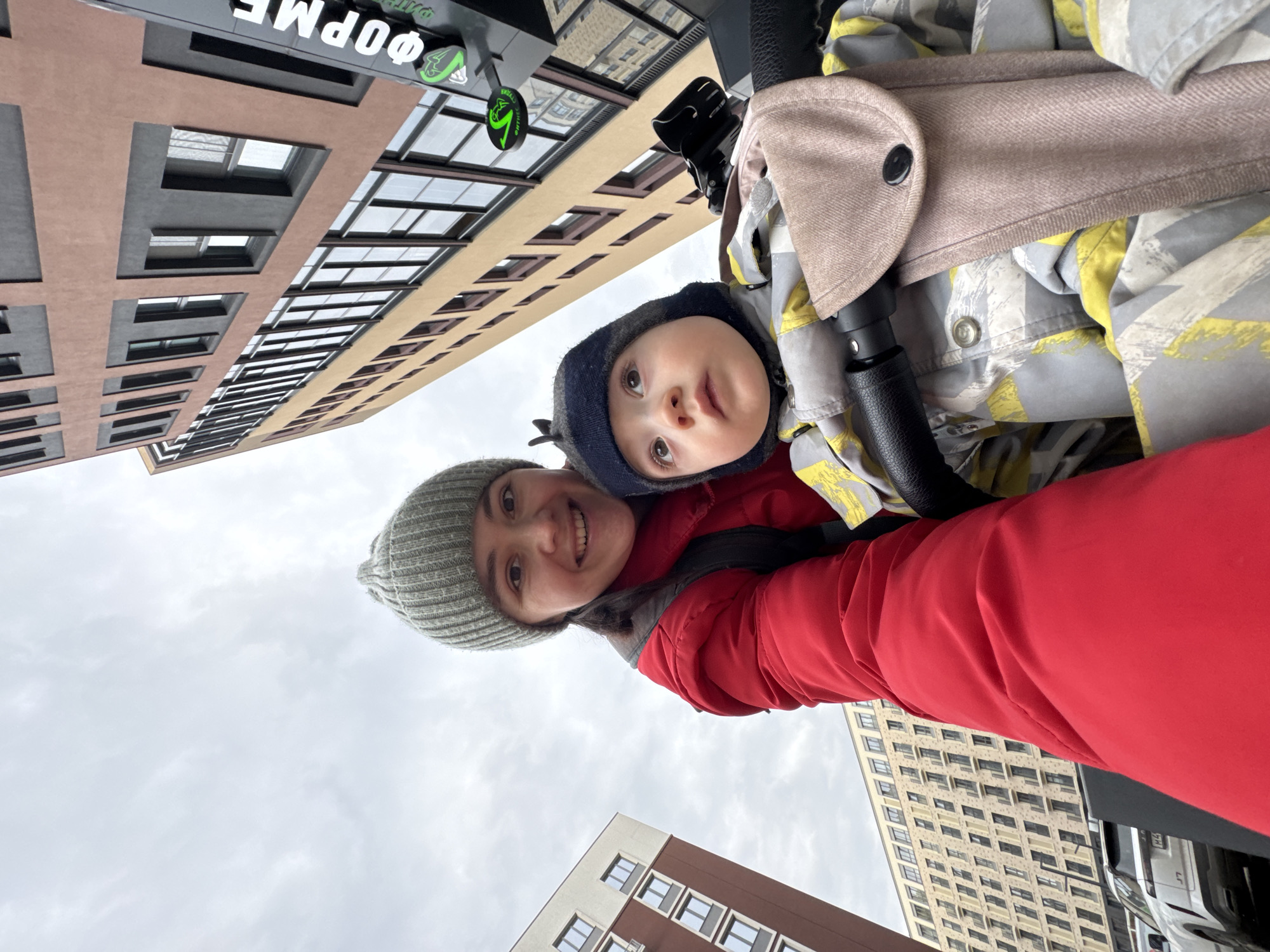 A low-angle portrait shows a woman wearing a gray knitted hat and a red jacket, smiling with a baby in a gray and yellow patterned hooded jacket. The baby is in a stroller. In the background are tall buildings. One building has a light brown and gray facade, and another a tan facade. A sign with Cyrillic text and a green logo is visible. The location is Izhevsk, Russia.