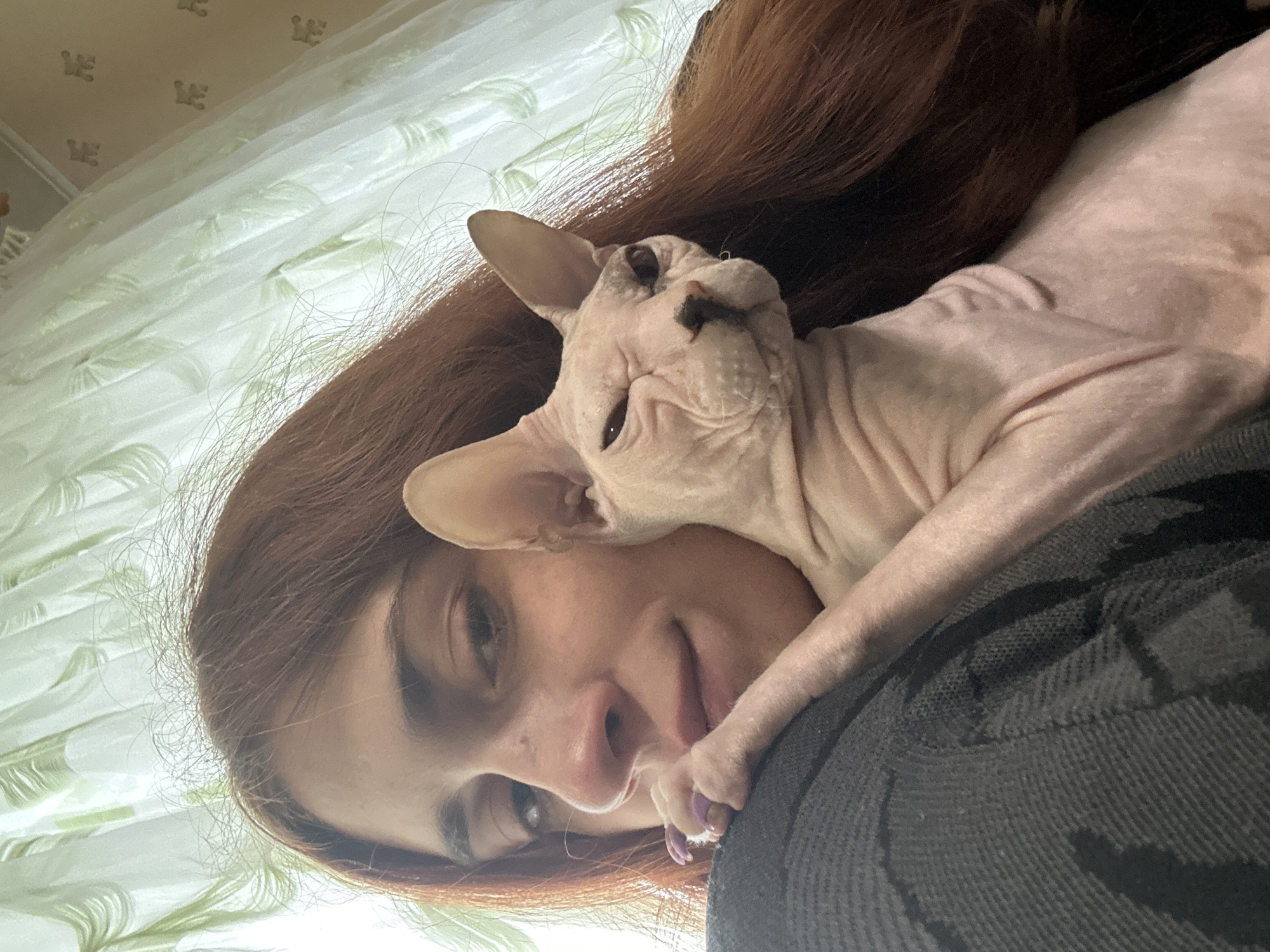 This is a close-up, vertically oriented selfie featuring a person and a Sphynx cat. The person, with reddish-brown hair, is looking up and slightly to the side, with their face positioned close to the camera. Their eyes are open and seem to be looking at the camera. The Sphynx cat, with its characteristic wrinkled pink skin, is resting on the person, with its head nestled near the person's cheek. The cat's eyes are partially closed, and its mouth is slightly open, revealing a pink tongue. One of the cat's paws is resting on the person's chest, with its claws extended. The background consists of a sheer curtain with a subtle green pattern and a wall with a patterned wallpaper. The lighting suggests it might be daytime, with natural light coming from the side, illuminating the subjects and creating soft shadows. The overall mood appears intimate and relaxed, with the person and the cat seemingly comfortable and affectionate with each other. No text is visible in the image. The provided location context is Togliatti, Russia.