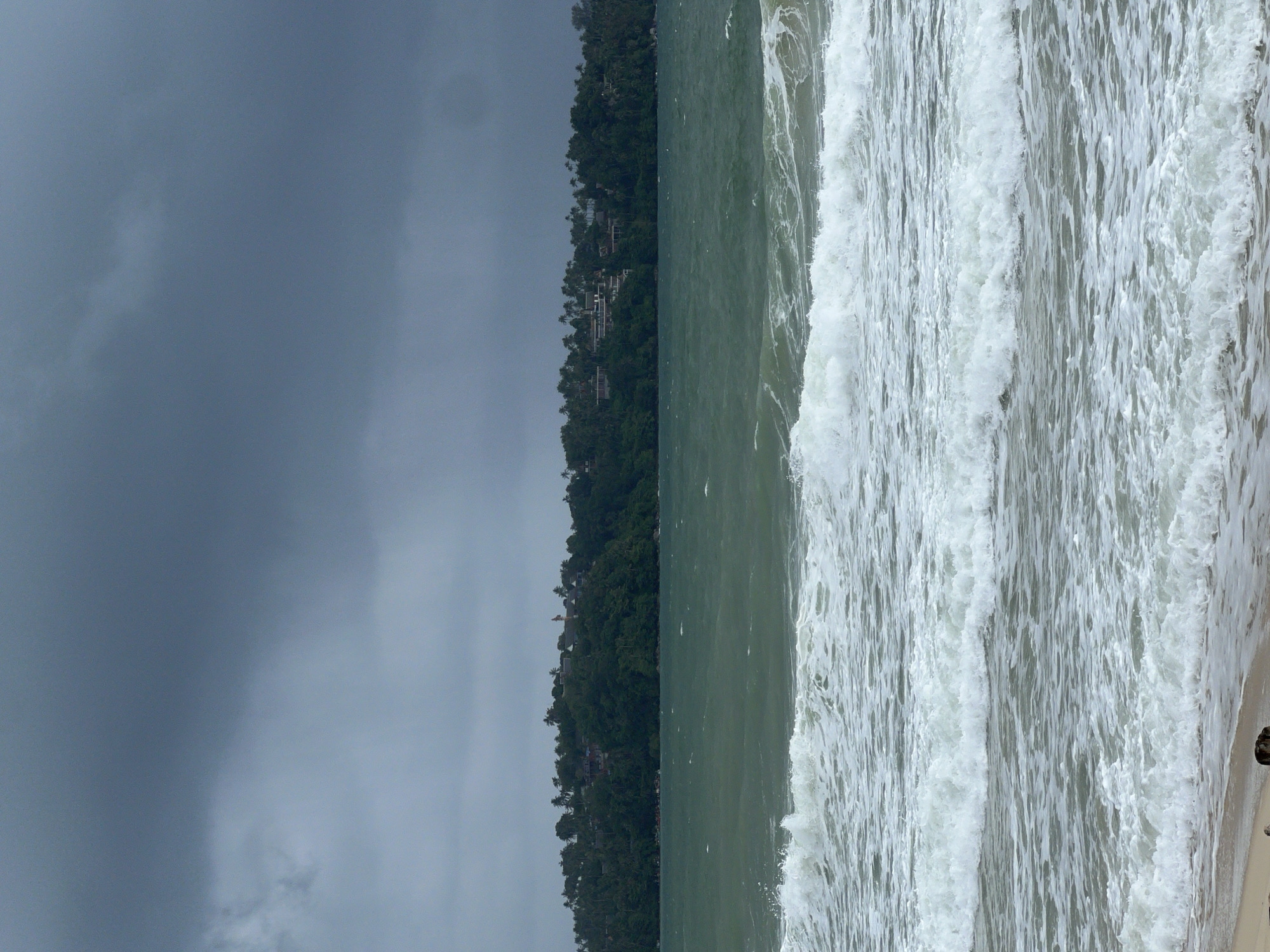 The image captures a dynamic coastal landscape under a heavily overcast sky. The upper left portion of the frame is dominated by a dense, dark grey-blue sky, indicating cloudy or stormy weather. Below the sky, a prominent landmass stretches horizontally across the middle of the image, densely covered with lush, dark green tropical vegetation, likely trees or jungle. Several structures, possibly houses or resort buildings, with light-colored roofs are visible nestled within this hillside foliage.
The right side of the image features the ocean, characterized by its deep, somewhat murky greenish-grey water. Numerous vigorous, white-capped waves are actively crashing and rolling towards the shore, creating a turbulent and foamy surface across the water. A sliver of sandy beach is discernible at the very bottom right corner, partially obscured by the incoming waves. No people or additional distinct objects are visible. The scene portrays a powerful natural environment, possibly during rough seas or before/after rain, under the dim light of an overcast day. There is no visible text.