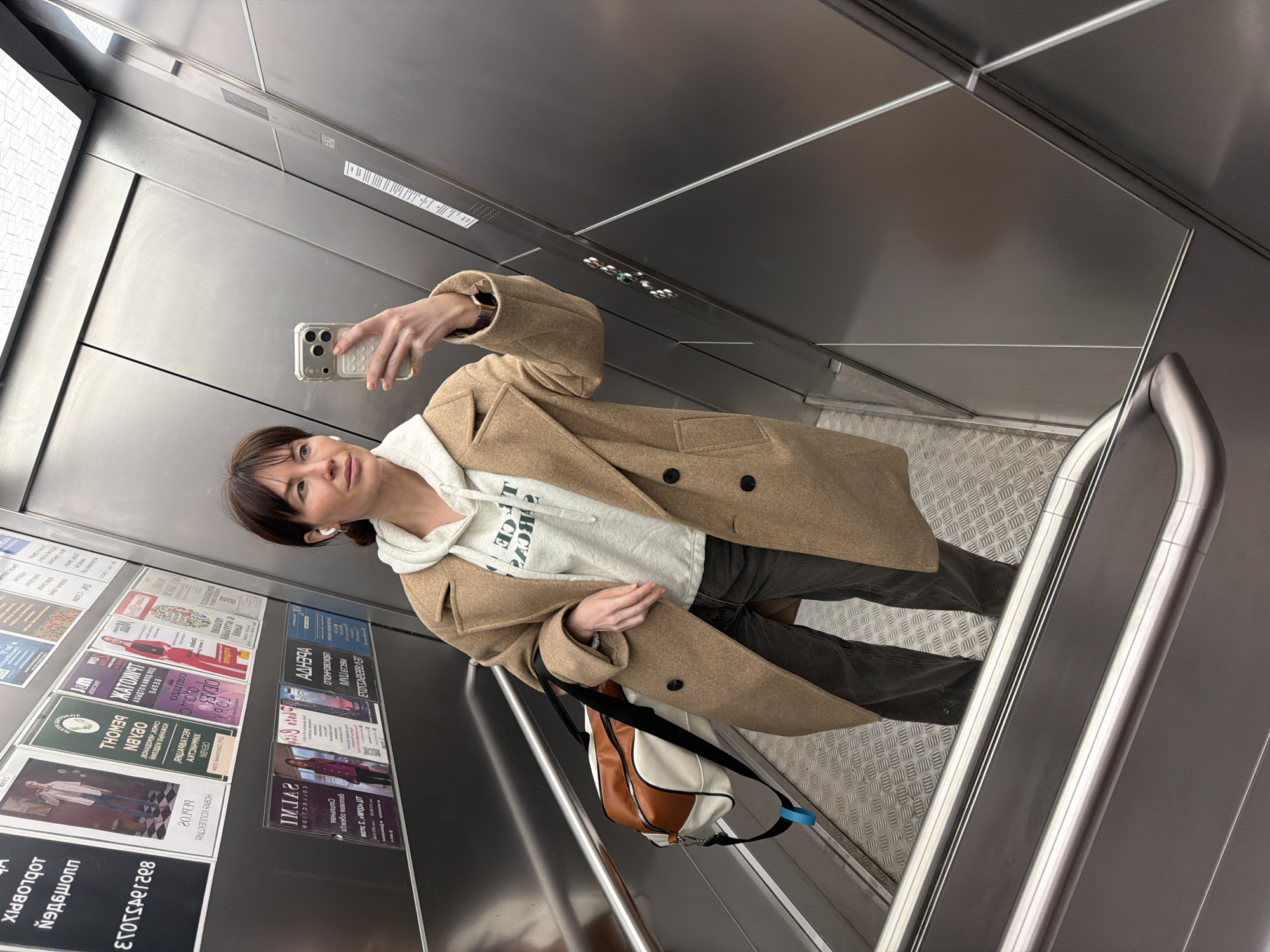 A woman is taking a selfie in what appears to be an elevator. She is wearing a tan overcoat, a grey hoodie with green lettering, and dark pants. She has a shoulder bag hanging from her shoulder. The elevator has a stainless steel interior. On one of the elevator walls, there are numerous posters and advertisements, many of which appear to be in Russian and mention "Perm". The floor of the elevator is a silver textured metal, possibly a tread plate. A stainless steel handrail runs along one side of the elevator.