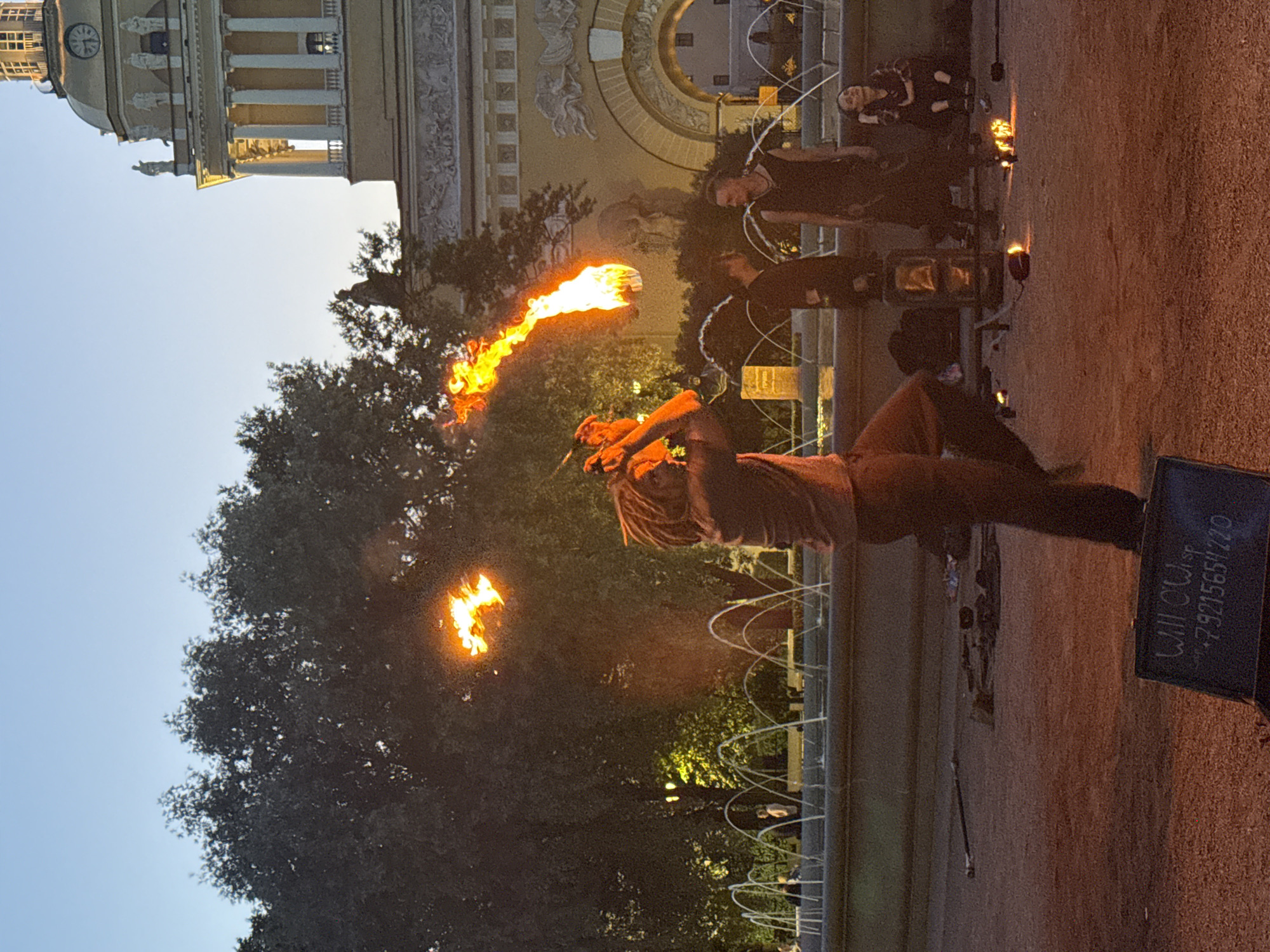 The image depicts an outdoor scene in Saint Petersburg, Russia, likely in the evening given the lighting and clear sky. In the foreground, a fire performer with dreadlocks is actively engaged in a fire-spinning act, with a ball of flame visible in mid-air. To the right, several spectators are observing the performance, with one individual sitting and looking upwards. Behind the performer and spectators is a fountain with multiple jets of water spraying upwards. In the background, a large, ornate building with classical architecture, including columns and statues, is visible. Trees with dark foliage are present on the left side of the image, and the ground is a gravelly, reddish-brown surface. A sign with writing is visible in the bottom left corner of the image.