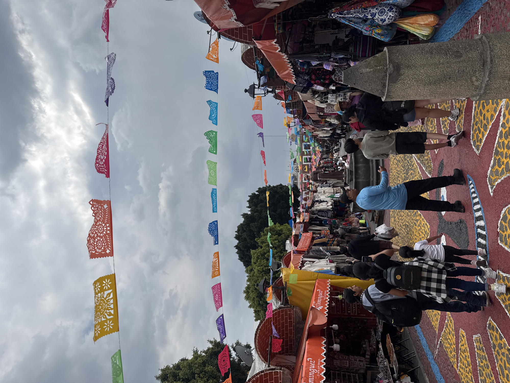 The image captures a street scene in Puebla, Mexico, bustling with people walking along a decorated street. Colorful papel picado banners hang overhead, stretching across the street between market stalls. These stalls are lined with merchandise, suggesting a market or fair setting. People are the primary subjects of the photo, walking in both directions along the street. The ground is painted with an intricate pattern. The sky is overcast.