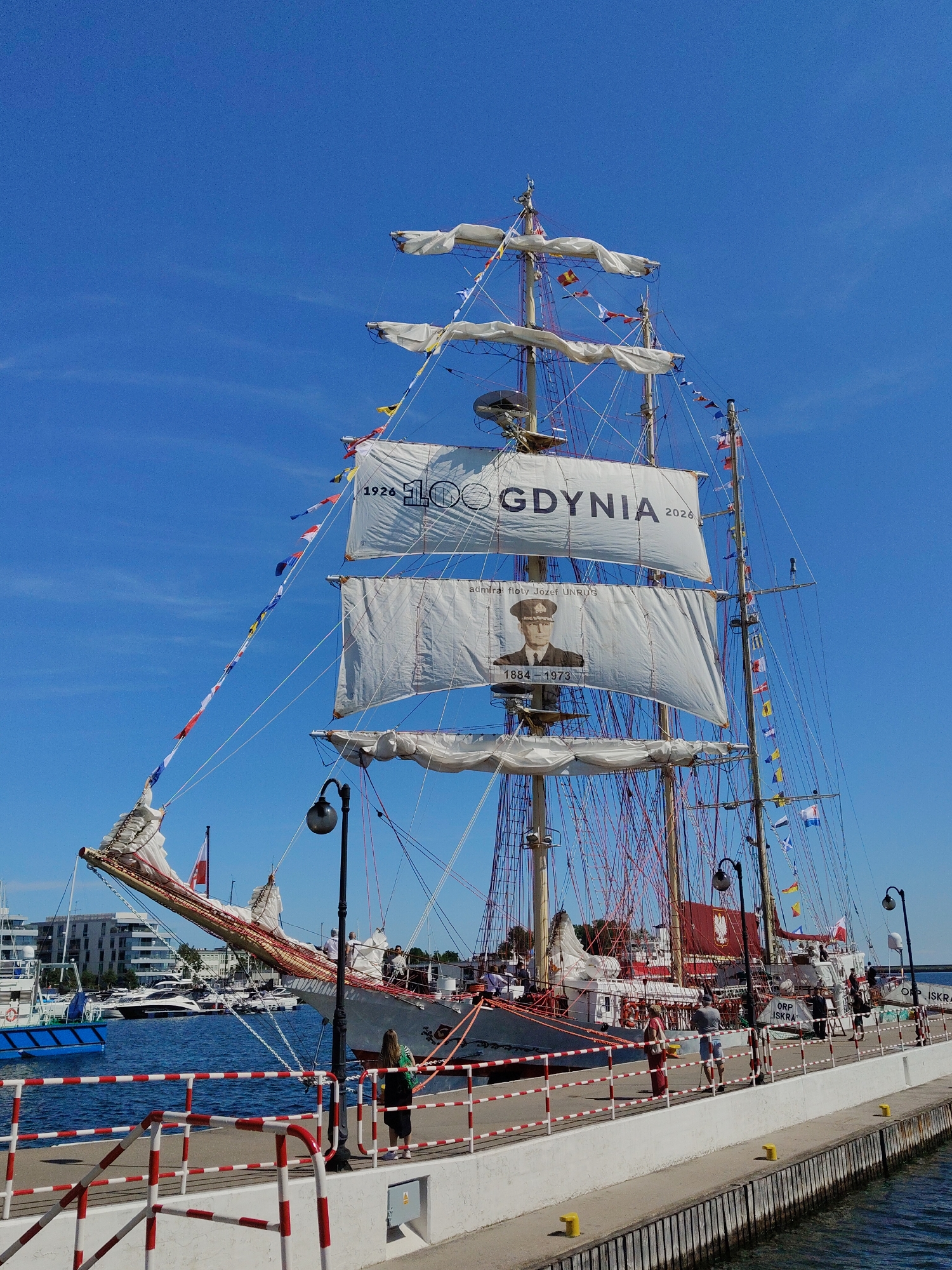 The image depicts a large sailing ship docked in the city of Gdynia, Poland, under a clear blue sky. The ship, identified as ORP ISKRA, is adorned with white sails, some bearing inscriptions. One sail displays "1926 100 GDYNIA 2026", indicating a centenary celebration. Another sail features a portrait of a man in military attire, with dates "1884-1973" below. The ship is also decorated with numerous colorful flags and pennants strung between its masts.
In the foreground, a white concrete pier with red and white striped railings runs along the waterfront. Several people are visible on the pier, observing the ship and its surroundings. A woman in a green top and black skirt stands near the railing, looking towards the water. A man in a grey t-shirt and blue shorts walks with a cane. Another woman in a red dress is also present. In the background, other boats and modern buildings can be seen, suggesting a bustling harbor or marina. The water is a deep blue, reflecting the sky. The overall scene suggests a public event or celebration related to the ship or the city's maritime heritage.