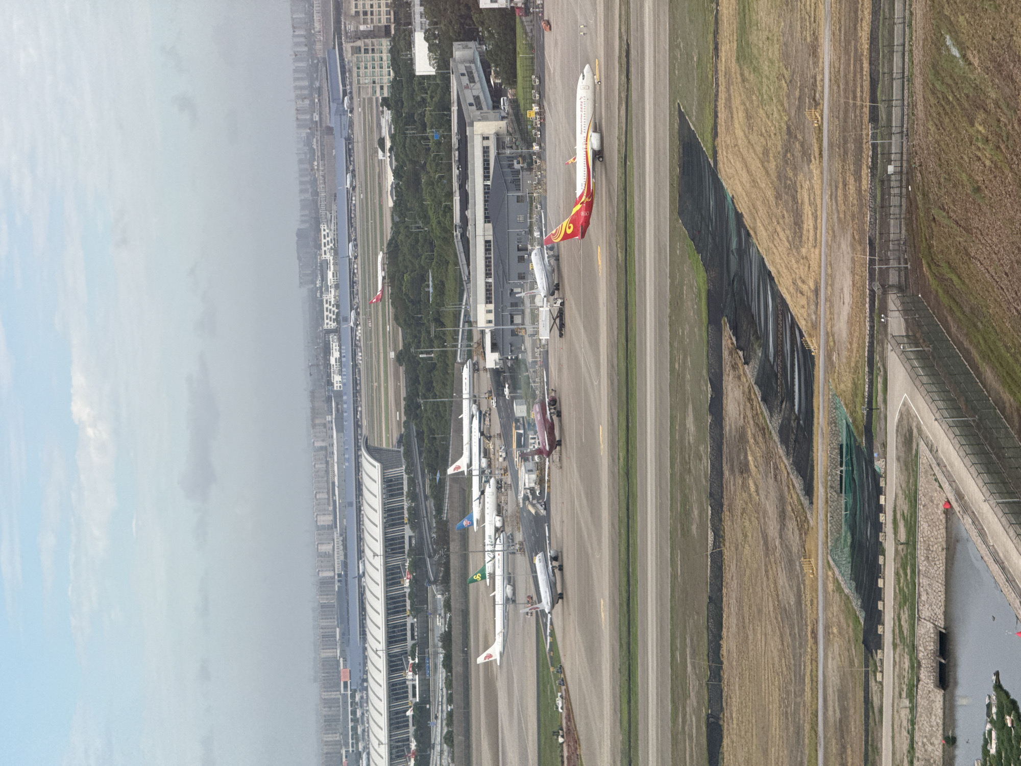 This is an aerial photograph of a busy airport tarmac in Pudong, China, likely Shanghai Pudong International Airport, given the context of Pudong being a major hub. The image captures multiple commercial airplanes parked and taxiing on the asphalt runways and taxiways. A large, modern airport terminal building with a curved roof is visible in the background, with a cityscape stretching out beyond it. The foreground shows undeveloped land with patches of dry grass and some construction or erosion control measures in place, including netting and stone-lined ditches. The sky is overcast, suggesting a cloudy or hazy day. Several vehicles, possibly service equipment, are scattered around the aircraft. The overall scene depicts a functional airport environment with ongoing aviation activity.