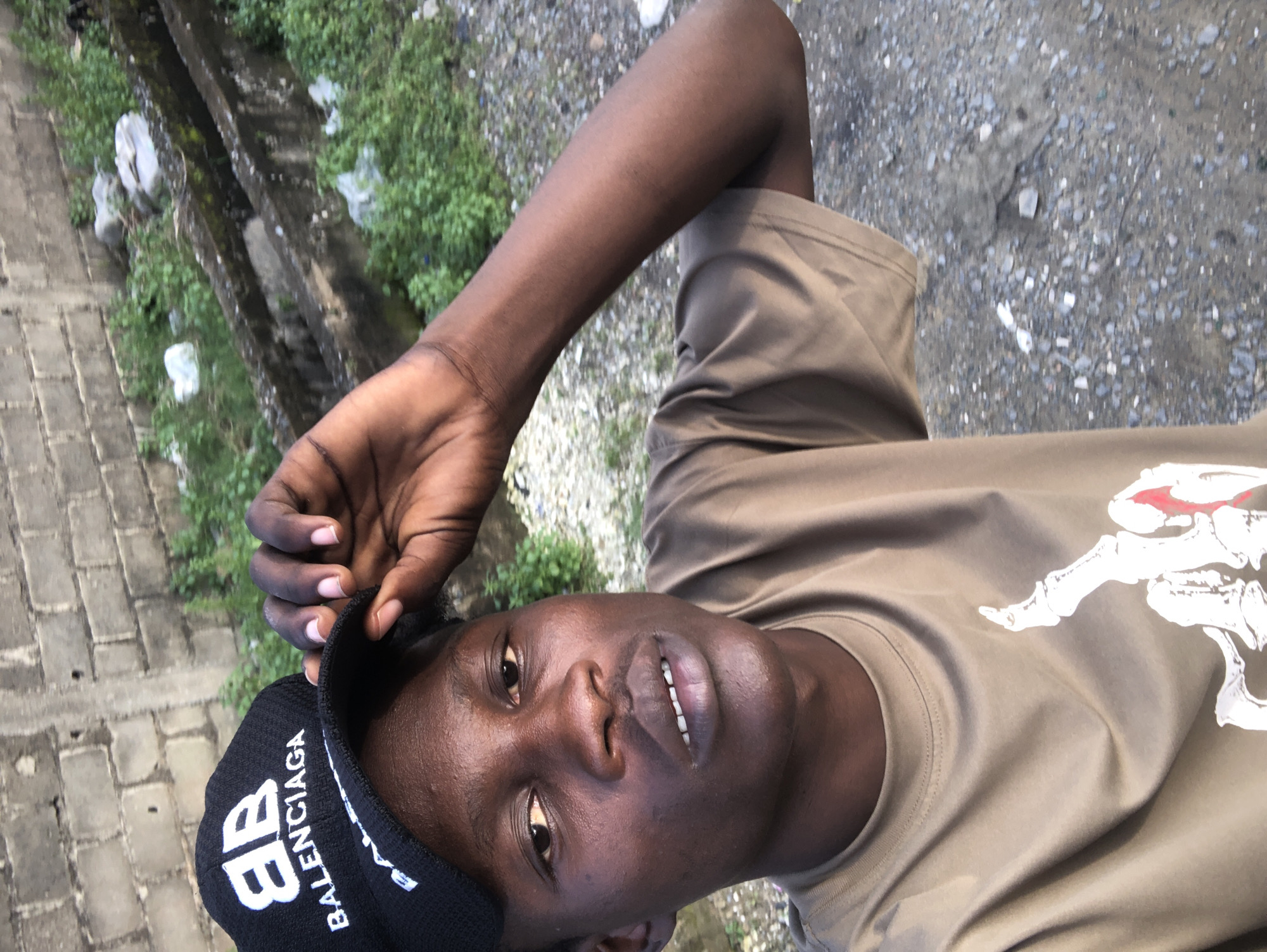 A young Black male is pictured outdoors, taking a selfie. He is wearing a black Balenciaga baseball cap with a white logo and a tan t-shirt with a white graphic. His left hand is on the brim of his cap, adjusting it. The background consists of a brick wall, some overgrown green vegetation, a concrete structure that appears to be a drain or channel, and gravelly ground. The overall environment suggests an urban or semi-urban setting.