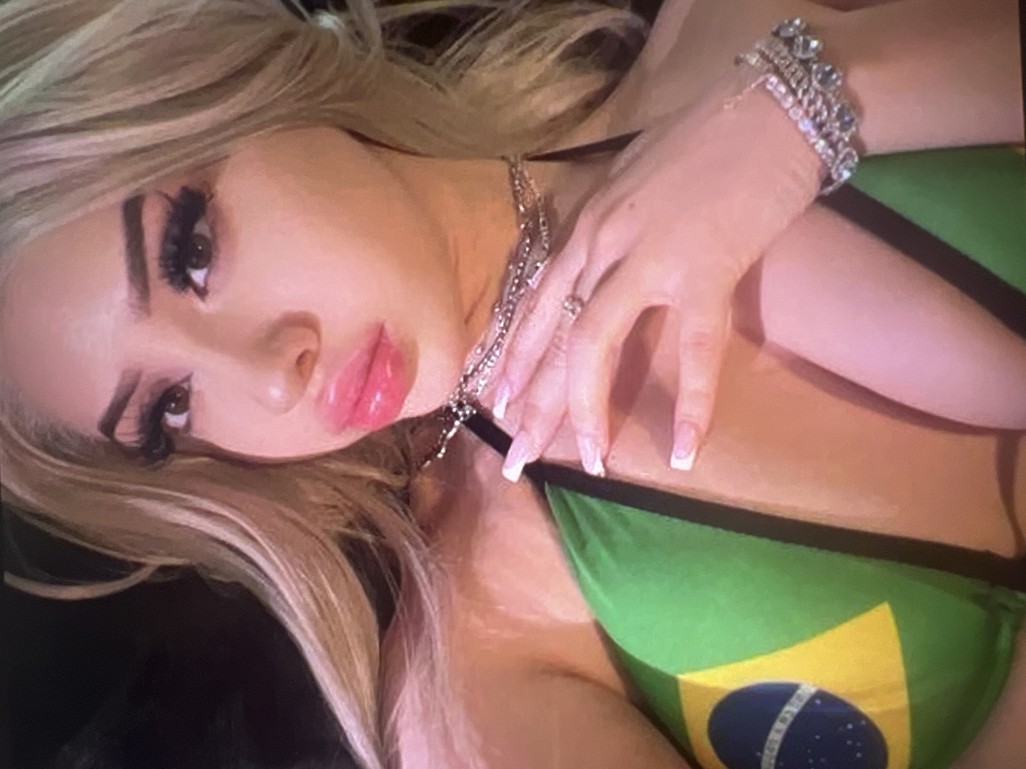 A woman with blonde hair and prominent makeup is shown in a close-up shot, wearing a bikini top that features the Brazilian flag. She has large, full lips with glossy pink lipstick, dramatic false eyelashes, and is adorned with several silver necklaces and a diamond bracelet. Her right hand, with manicured nails, rests on her chest. The background is dark and out of focus. The image provides no information about the city of Minsk, Belarus.