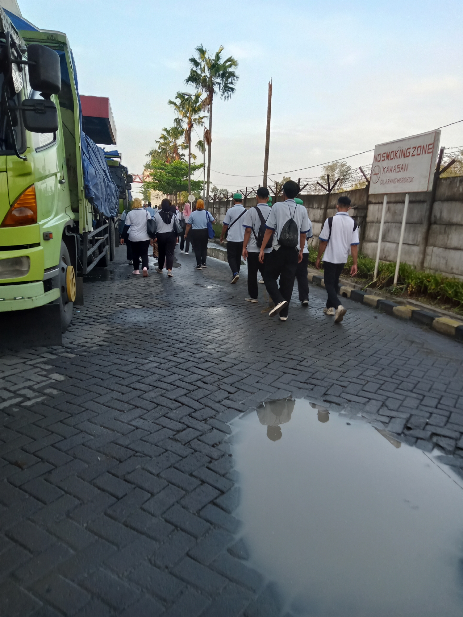 A group of people are walking on a brick road in Sukomoro, Indonesia. A green truck is parked on the left side of the road. On the right side of the road is a concrete wall with barbed wire on top and a sign that reads "NO SMOKING ZONE." A puddle is visible on the road. Palm trees are visible in the background.