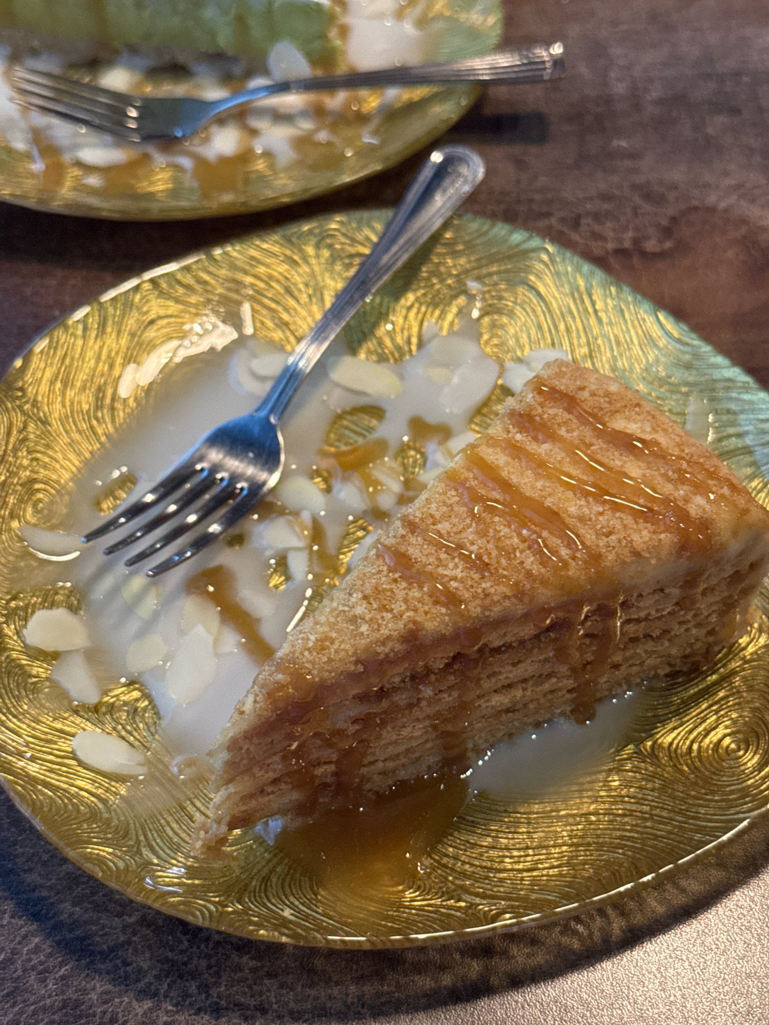 A slice of layered cake with caramel drizzle and slivered almonds sits on a decorative gold plate. A fork rests in the sauce on the plate. In the background, another similar plate with a fork and a portion of cake is visible. The table surface is a dark, textured material. The scene is photographed from a slightly elevated angle. The image does not provide any information about its location in the city of Chaikovsky, Russia.