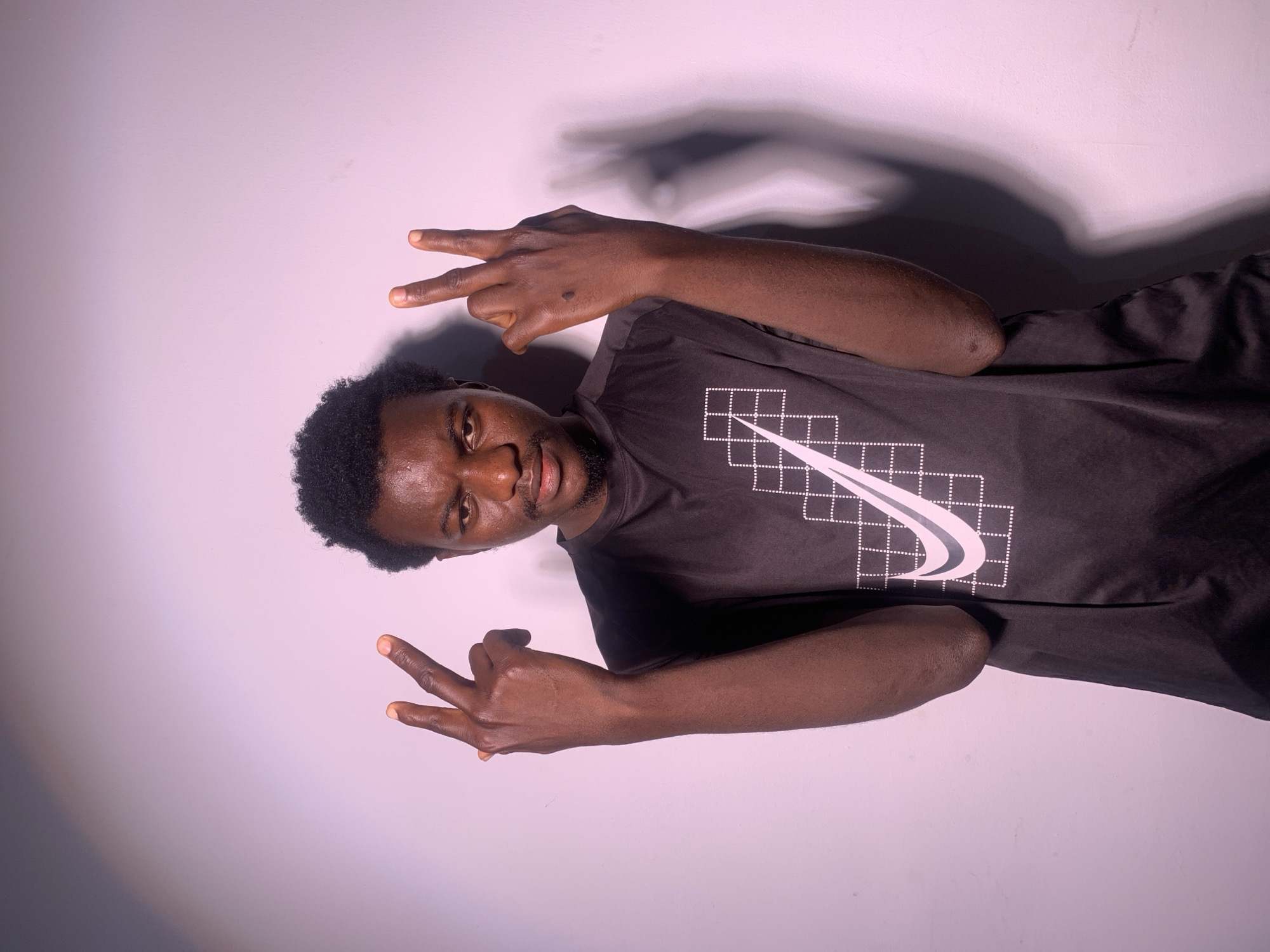 A dark-skinned man is shown in a vertical, close-up, medium shot against a light-colored wall. He is wearing a black t-shirt with a white Nike logo and a grid pattern. The man has dark, curly hair and a short beard. His head is tilted to the side, and he is looking directly at the camera with a neutral expression. His right hand is raised, and he is holding up two fingers in a peace sign. His left hand is also raised and making the same gesture. Shadows are cast on the wall behind him, suggesting a light source from the side. The image appears to have been taken indoors, possibly in a studio, due to the uniform background and controlled lighting. No specific time of day or weather conditions are evident. The overall mood is casual and posed.