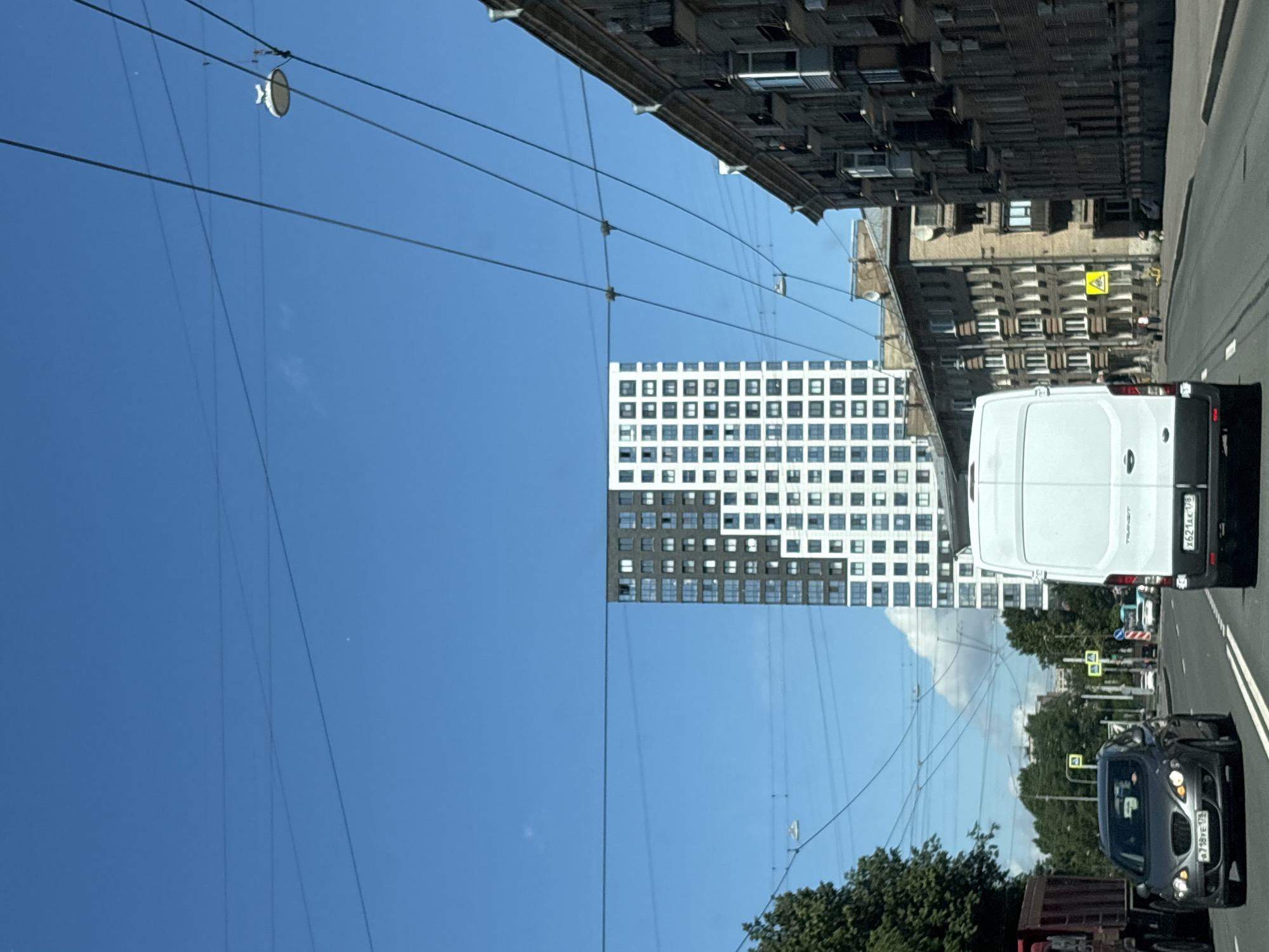 The image depicts a street scene in Saint Petersburg, Russia, with a modern high-rise building contrasting with older architectural styles. In the foreground, a white transit van is driving on a road, followed by a dark-colored sedan. Overhead, a network of trolleybus wires crisscrosses the sky, with a single streetlamp visible attached to them. To the right, a multi-story brick building with many windows and balconies lines the street, showcasing older architecture. On the left side of the image, a tall, contemporary apartment building with a checkerboard pattern of dark and white facades stands out against the clear blue sky. Trees and smaller buildings are visible in the background, suggesting a mix of residential and commercial areas within the city. The scene appears to be captured during daylight with sunny weather.