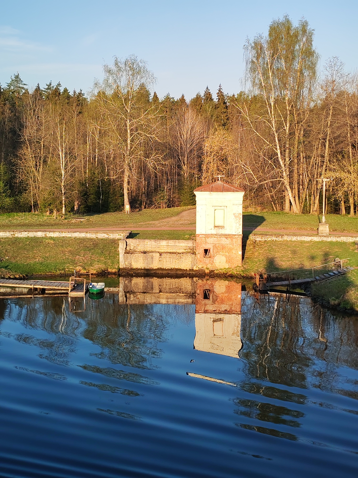 The image shows a small, old brick building with a light-colored, possibly concrete, top section and a cross on its roof. It stands on the edge of a body of water, with its reflection visible in the water. Adjacent to the building are concrete retaining walls. A small wooden dock with a green rowboat is located on the left side of the building.
Behind the building is a grassy area with trees in the background. A lamp post is visible near the right side of the building.
The location is likely in Zaprudnya, Russia, based on the description provided in the prompt.