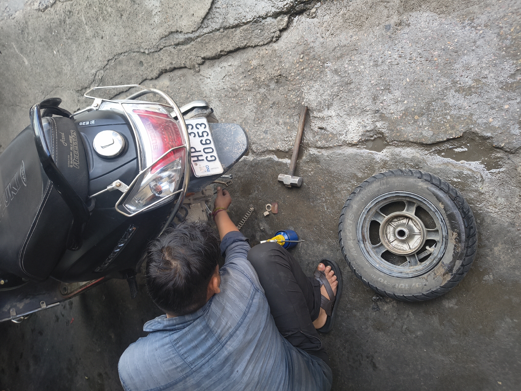A man is working on a dark-colored scooter in what appears to be an outdoor workshop or a roadside repair area. He is wearing a light blue striped shirt and dark pants, with brown sandals on his feet. The man is bent over, focusing on the rear wheel area of the scooter, which has been removed and lies on the ground beside him. A tire is visible, with "MAXXIS" branding. Tools, including a hammer and a yellow oil dispenser, are scattered on the wet, cracked concrete ground. The scooter has a license plate that reads "HP 33 G0653". The overall scene suggests a repair or maintenance activity taking place in an urban or semi-urban environment, potentially in Dharamshala, India, given the license plate. The ground is wet, indicating recent rain or water usage.