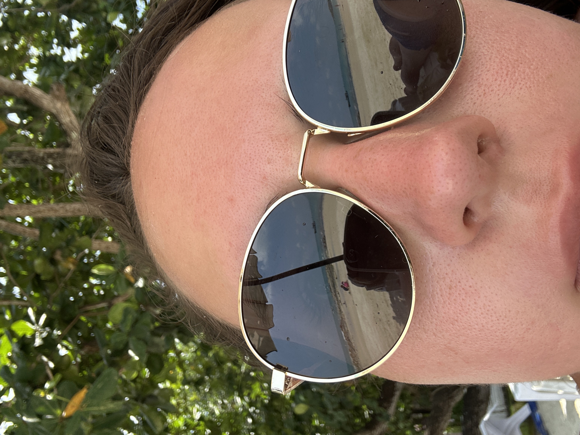 Close-up perspective of a person's face wearing gold-rimmed sunglasses, revealing a beach scene reflection in the lenses. The background includes green foliage. The location is identified by the beach scene reflection, indicating Ao Noi Na, Thailand.