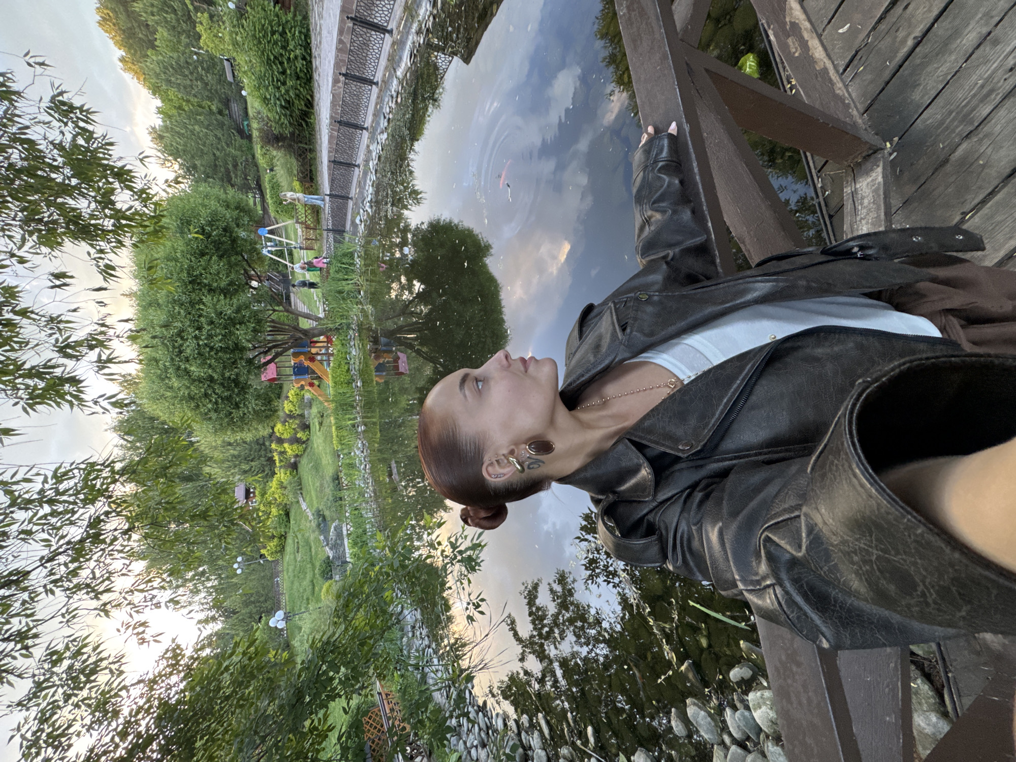 The media file displays a serene outdoor scene, possibly a park, in Izhevsk, Russia. The primary subject is a young woman, seen from the chest up, positioned on the right side of the frame. She is leaning against a rustic wooden railing or bridge, looking upwards and to the right, with a contemplative expression. Her hair is styled in a neat bun, revealing a distinctive large, gold-toned earring with a darker insert, and smaller ear piercings. She wears a black leather jacket over a white top, and a delicate gold chain necklace is visible. Her fingernails are painted a light color.
In the midground, a calm body of water, likely a pond or river, reflects the sky, showing soft clouds and hints of bright light. A fish, possibly a Koi, is visible in the water, creating ripples. The wooden structure the woman is leaning on features rough-hewn planks. Along the water's edge in the foreground are various sized smooth rocks.
The background reveals a lush green landscape. Across the water, there's a paved pathway lined with ornamental metal fencing. Beyond this, a grassy area dotted with trees and shrubs stretches into the distance. A colorful children's playground with swings and slides is visible. In the distance, near the playground, another adult person and a few children are present, appearing small in the frame. The overall lighting suggests it is late afternoon or early evening, with a soft, natural glow filtering through the trees. No visible text is present in the image.