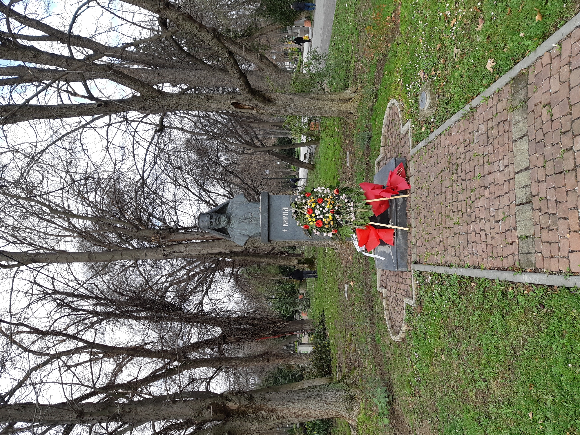 The image displays a bronze bust of Patriarch Kiril of Bulgaria, situated on a dark stone pedestal within Tsarevets Garden (also known as the City Garden) in Plovdiv, Bulgaria. The bust depicts a bearded male figure wearing ecclesiastical vestments, including a religious head covering resembling a mitre and a pectoral cross. The name "+ КИРИЛ" is inscribed in Cyrillic on the pedestal. A large commemorative wreath, featuring red, white, and yellow flowers with prominent red ribbons, rests against the front base of the monument.
The monument is positioned on a patch of green grass and is approached by a reddish-brown cobblestone pathway. The surrounding environment consists of a public park characterized by numerous mature deciduous trees, largely bare of foliage, indicating a cold season. The sky above is overcast. In the background, a paved pathway extends through the park, populated by several individuals. One person in dark attire stands facing away from the camera on the left side of the scene, while others are observed walking casually along the path on the right, one carrying a yellow bag. No direct interactions between the individuals are discernible. The scene is static, focused on the memorial in a tranquil park setting.