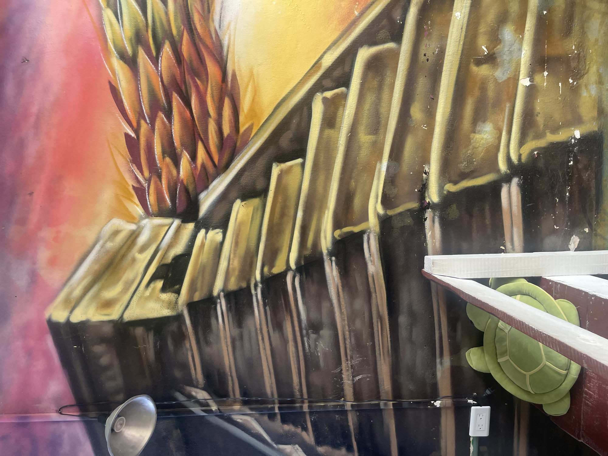 The image depicts a section of a mural, possibly in Playa del Carmen, Mexico. The mural features a stylized, oversized pineapple and a series of stacked, rectangular shapes, rendered in gold and dark brown hues. In front of the mural, there is a metal lamp, a green plush turtle, and a wooden structure that could be a ladder or shelf. A white electrical outlet is visible near the base of the structure. The wall itself shows signs of wear and tear, with scuffs and paint imperfections.