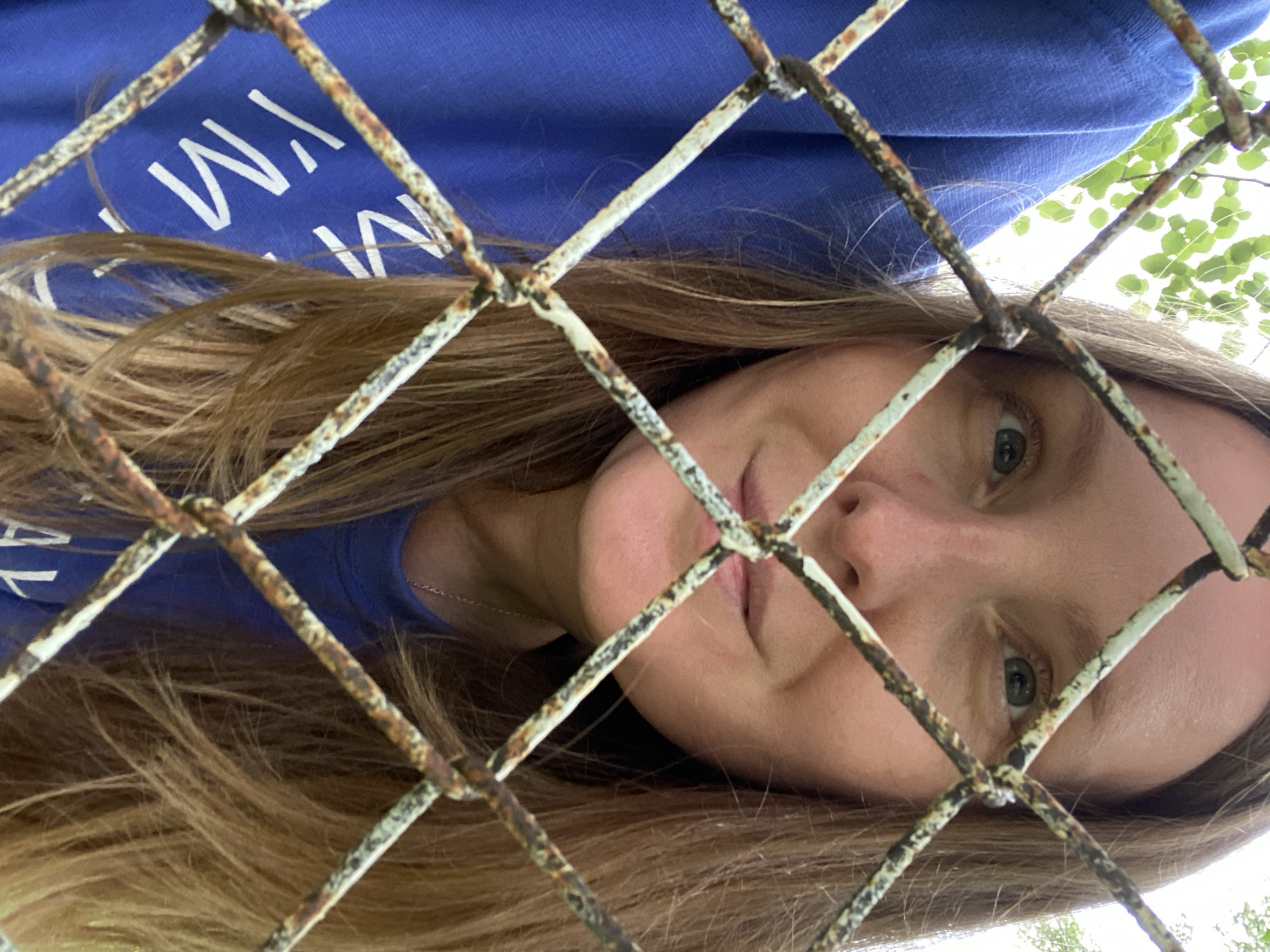 This is a close-up, low-angle selfie of a young woman taken through a rusty chain-link fence. She is wearing a bright blue hoodie with white text on it, which is partially visible. The text appears to say "I'M MY OWN..." with the rest obscured. Her long, light brown hair falls around her face and shoulders.
The woman's face is turned towards the camera, and she is looking directly into the lens. Her eyes are a striking green. The lighting suggests it is daytime, possibly late afternoon or early evening, as the light is somewhat diffused and there are shadows cast by the fence. The background shows green foliage, indicating an outdoor setting, likely in a park or a natural area, consistent with the location context of Noviy, Russia.
The mood of the photo is somewhat contemplative, with the fence acting as a visual barrier that adds a layer of introspection to the portrait. The woman's expression is neutral but engaging. The focus is sharp on her face and the fence, with the background slightly blurred. The rust on the fence is a prominent detail, adding texture and a sense of age or neglect to the foreground.