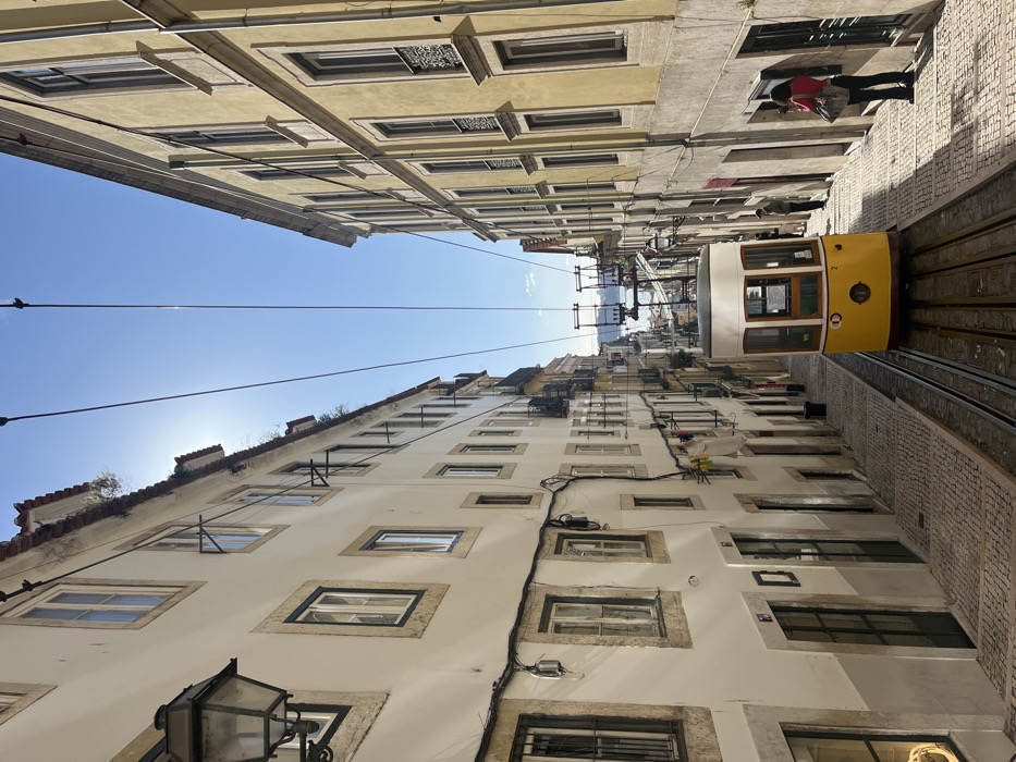 This image depicts the Rua da Bica de Duarte Belo in Lisbon, Portugal, with a prominent view looking downhill towards the Tagus River. A yellow, two-car funicular, marked with the number "2" on its front, is stationary and centrally positioned on its tracks in the lower half of the steep, cobblestone street. The street is flanked by multi-story buildings: the structure on the left has a cream-colored facade, while the building on the right exhibits a yellow facade. Both buildings feature numerous rectangular windows, some with small, dark-colored iron railings or frames. Overhead, multiple dark wires crisscross the clear blue sky, which shows a distinct sun flare at the top left. An ornate, dark-colored streetlamp is mounted on the upper left building. Utility lines run horizontally along the lower sections of the buildings, and a few pieces of white laundry are visible hanging from a window on the left side. On the right-hand cobblestone sidewalk, a person in a red top and dark trousers stands, holding a mobile device as if photographing the funicular or the view. Another individual is partially visible further down the right sidewalk.