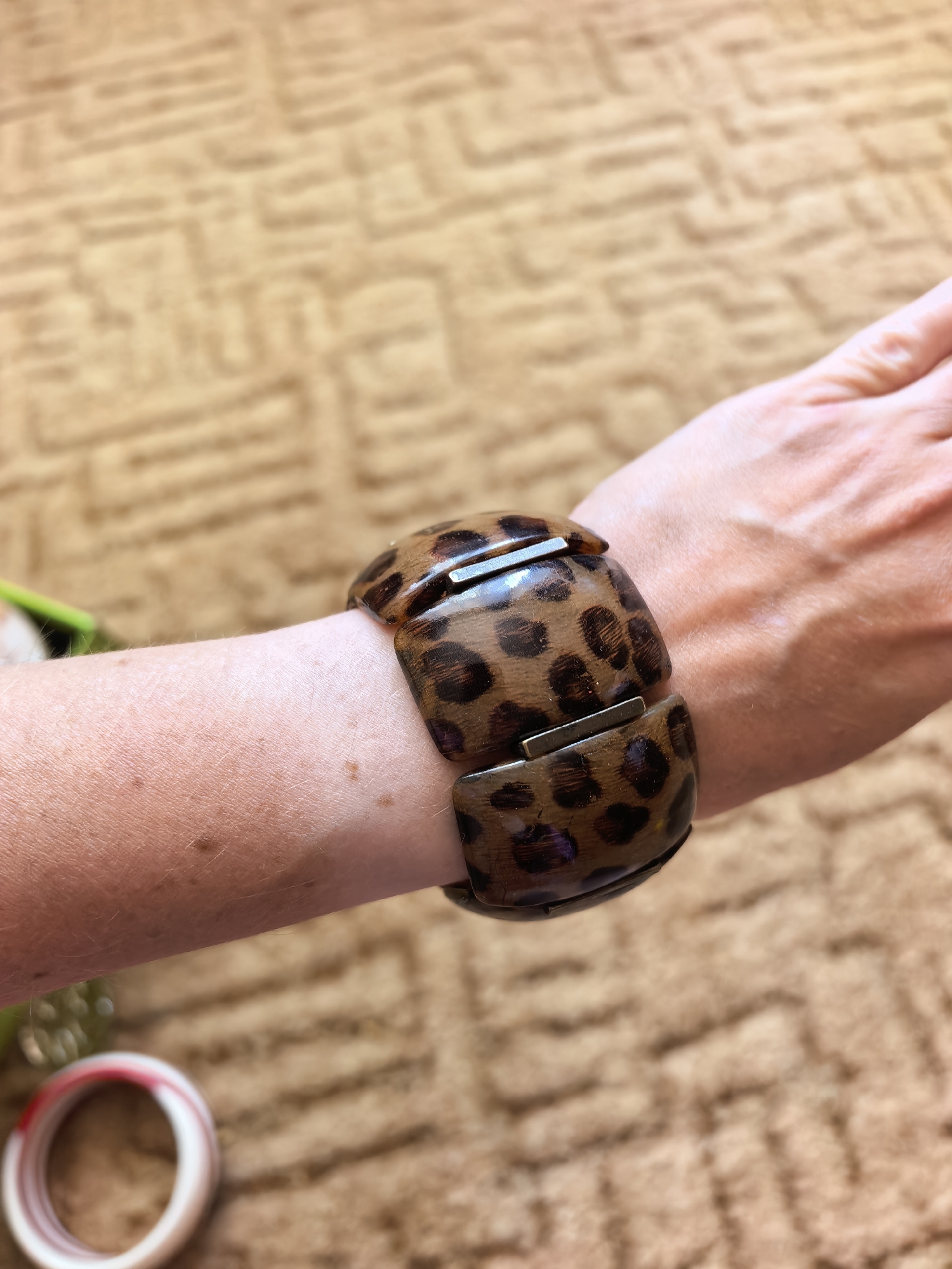 The image depicts a woman's forearm wearing a brown and black spotted bracelet, reminiscent of a leopard print, against a blurred brown patterned background. The bracelet appears to be made of segments connected by metallic bars. The woman's skin shows freckles and some visible veins. A portion of colorful bracelets is visible in the lower left corner of the picture. The image does not provide any information regarding the location in the city of Sterlitamak, Russia.
