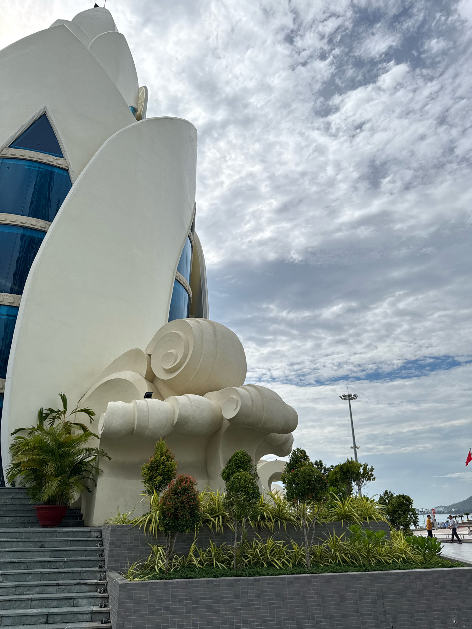 The image shows the exterior of the Nha Trang Institute of Oceanography, a building located in Nha Trang, Vietnam. The building features a unique, white, sculptural design resembling stylized petals or waves, with large blue-tinted windows. In the foreground, there are grey stone steps leading up to the building, with a large potted palm plant on the left. A well-maintained garden with various types of shrubs and plants is situated to the right of the steps, in front of the building's base. The garden is bordered by a grey tiled wall. In the background, a tall light pole stands near the water, and a red flag is visible on a pole further in the distance. The sky is partly cloudy. Two people are walking in the distance along a paved area by the water.