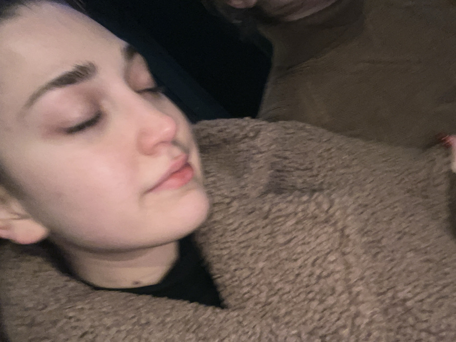A young woman with pale skin is asleep. Her eyes are closed, and her lips are slightly parted. She is wearing a black top and is draped in a brown, textured material resembling a fleece or fur coat. Part of another person is visible in the top right corner, wearing a similar brown garment. The background is dark.
