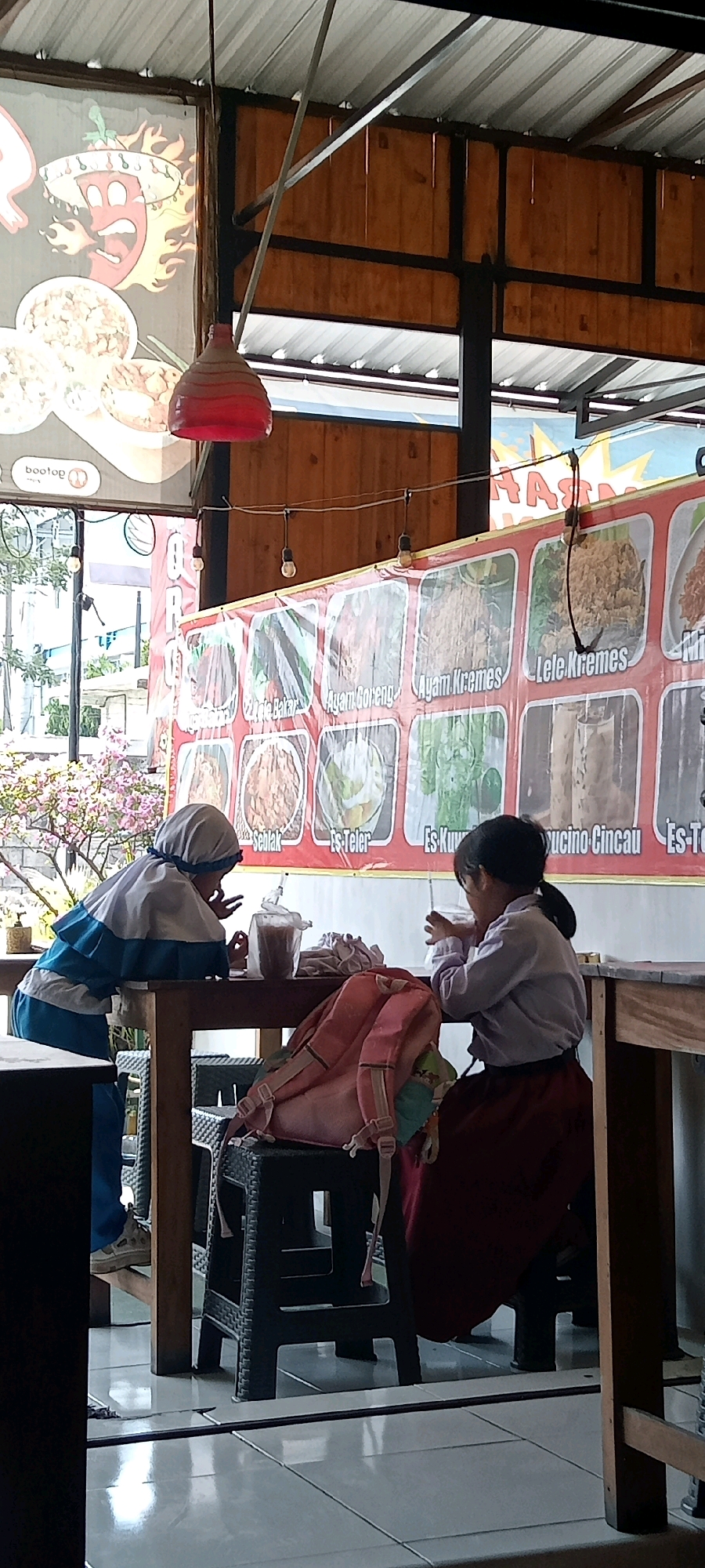 Two young females are seated at a wooden table under a covered outdoor dining area. The female on the left is wearing a blue and white hijab and jacket, and is leaning over the table, appearing to interact with something in front of her. The female on the right is wearing a purple shirt and a dark red skirt and is drinking from a cup. A pink backpack is placed on a black plastic chair between them. In the background, a large banner displays images of various food items, some with Indonesian text. The flooring is tiled, and the ceiling is corrugated metal with exposed wooden beams and metal framing. The scene appears to be a casual dining establishment in Pondok, Indonesia.