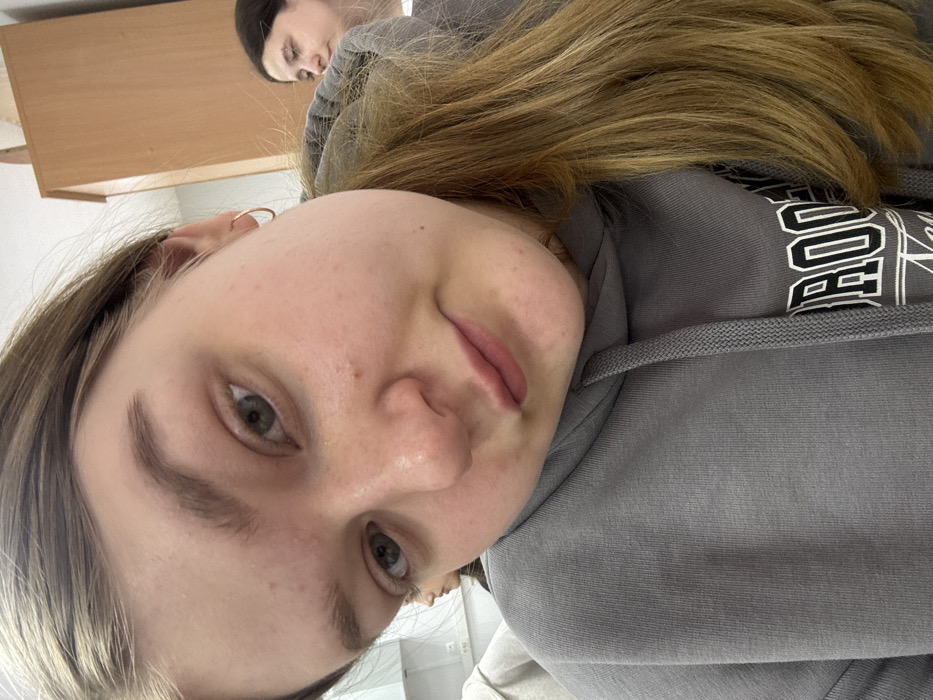The image depicts an indoor scene in Chaikovsky, Russia, within an environment consistent with a classroom or institutional building. No specific landmarks identifying the city are visible. The primary subject is a young female, appearing to be in her late teens or early twenties, positioned centrally in a close-up, selfie-style shot. She has light brown hair, light-colored eyes, and a slight pout on her lips as she looks towards the camera. She wears a grey hooded sweatshirt with a partially visible white graphic on the chest, reading "ROOKLYN NEW." A small gold hoop earring is present in her visible left ear. Behind and to the right of the primary subject, a second female with dark hair is partially visible, looking towards the right side of the frame. A third individual's head and shoulder, wearing a light-colored garment, are minimally visible behind the primary subject's left side. The background features light-colored walls, a white cabinet or shelving unit, and a white electrical outlet to the left, along with a segment of a light brown wooden cabinet or wardrobe on the right.
