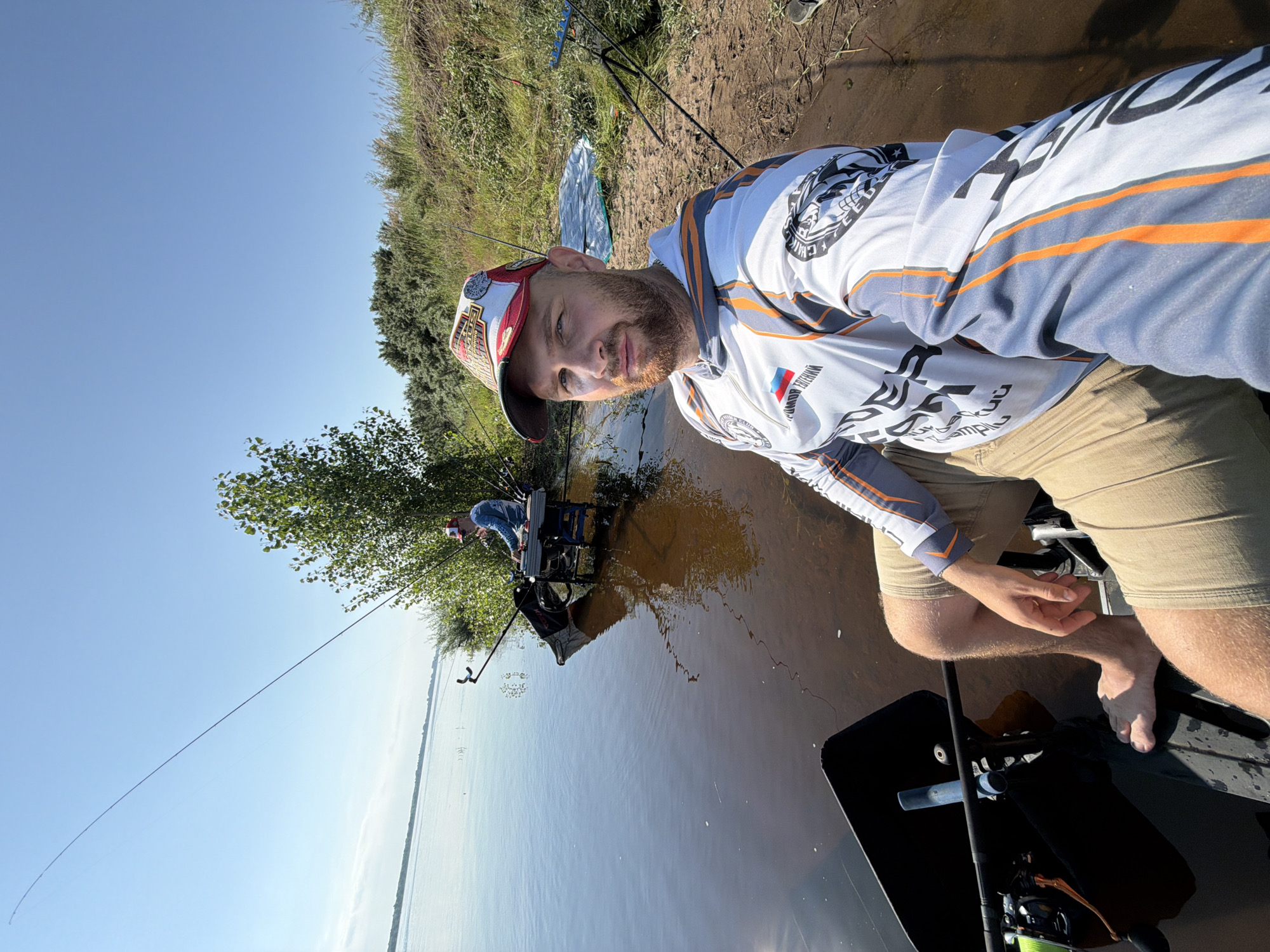 The image shows a man taking a selfie while fishing. He is sitting in a fishing seat on the bank of a wide river. The river is calm with some ripples and the water is dark brown. The bank is covered in dry grass and some sparse bushes. There is another person fishing further down the bank. The man in the foreground is wearing a fishing shirt with logos and a baseball cap. He has a beard and is looking at the camera. Fishing rods are visible in the foreground and background. The sky is clear and blue. The overall scene depicts a sunny day spent fishing outdoors.